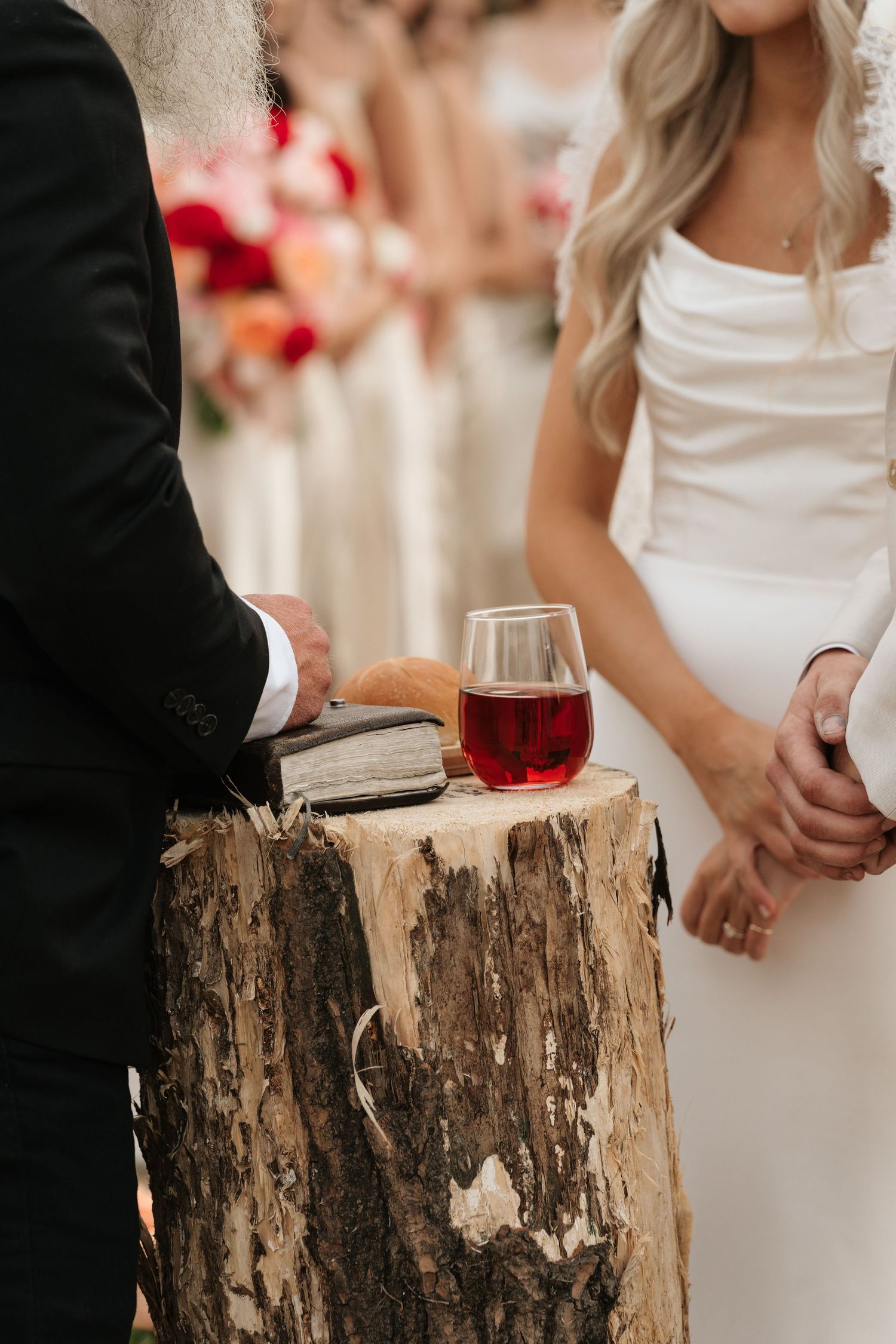 Wedding ceremony with red wine glass and Bible on rustic wood stump altar with bridal party in background.
