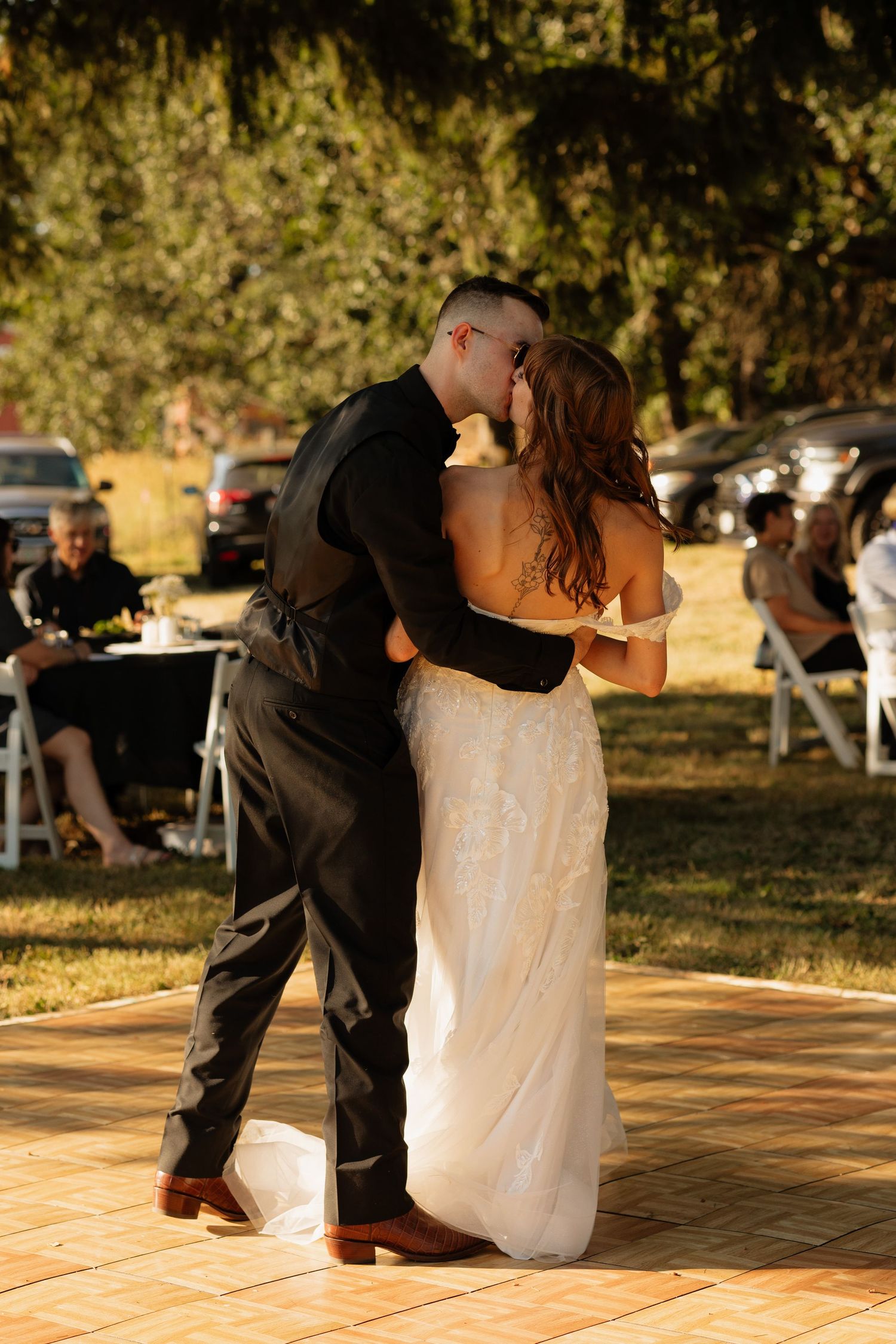 Bride and groom share a first dance kiss outdoors at a sunny wedding reception on a tiled dance floor.