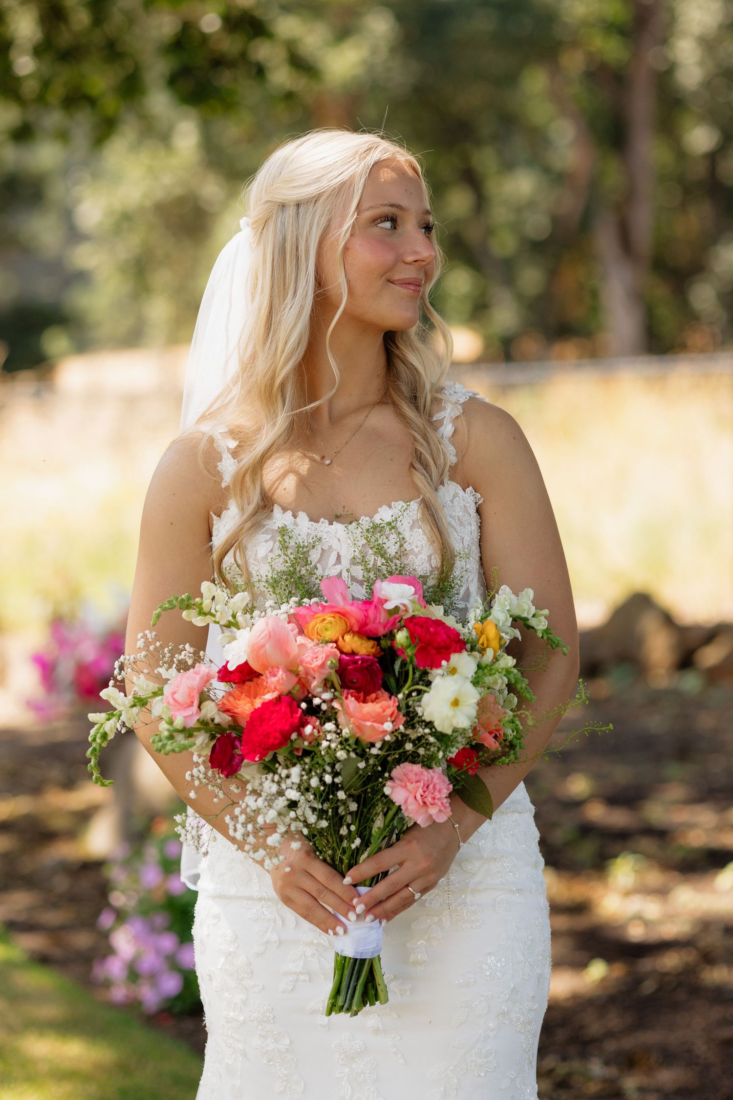Beautiful blonde bride in lace wedding gown holding colorful pink and red floral bouquet outdoors.