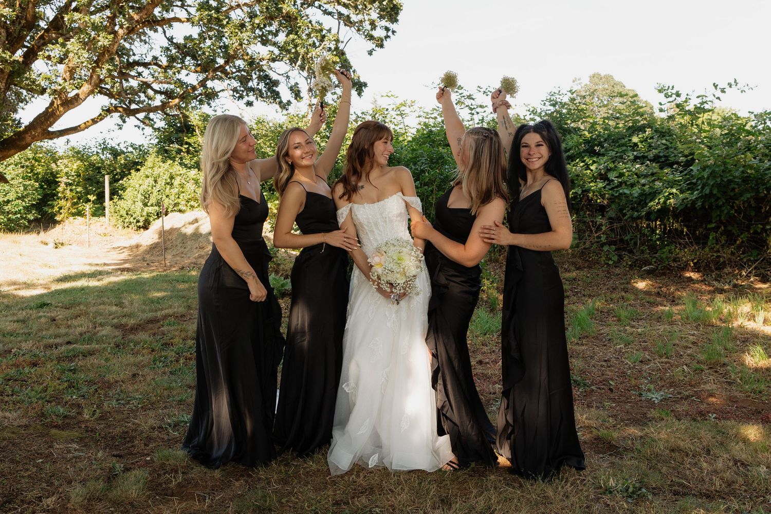 Bride in white gown with four bridesmaids in black dresses celebrating outdoors with raised arms and bouquets.