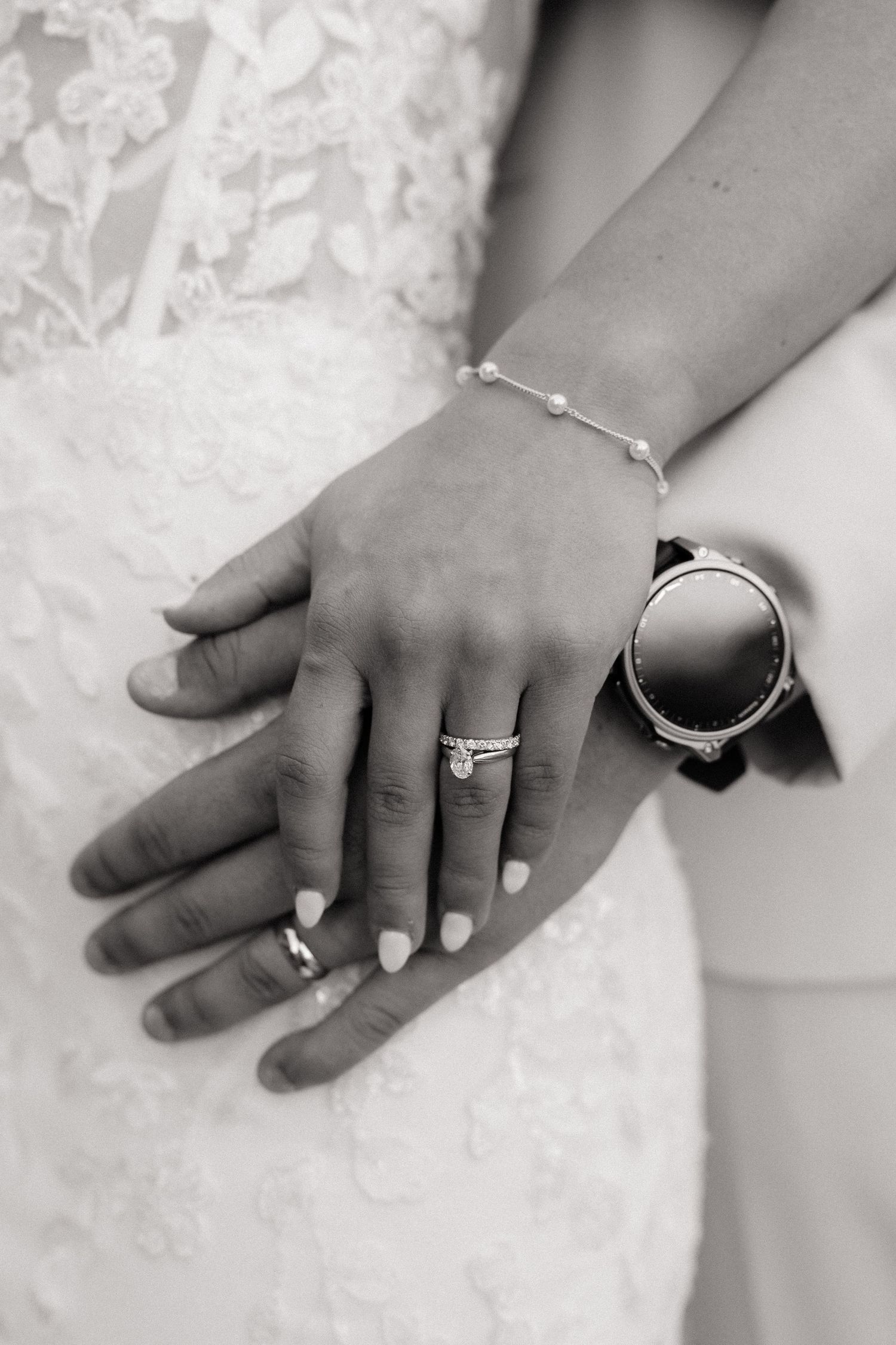 Black and white close-up of bride and groom hands showing wedding rings and pearl bracelet.