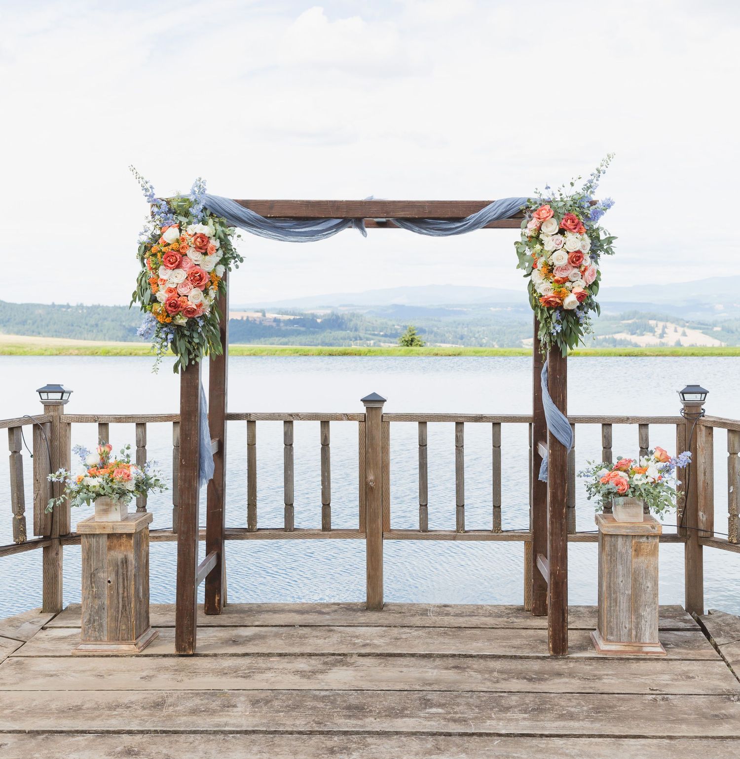 A rustic wooden wedding arch decorated with coral and peach floral arrangements overlooking a serene waterfront view.