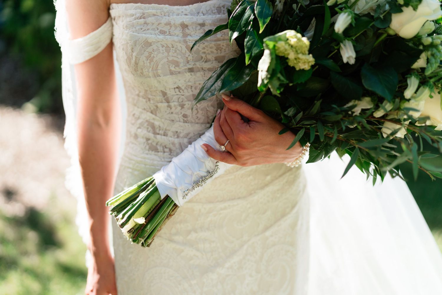 Bride in lace wedding dress holding elegant white and green floral bouquet outdoors.
