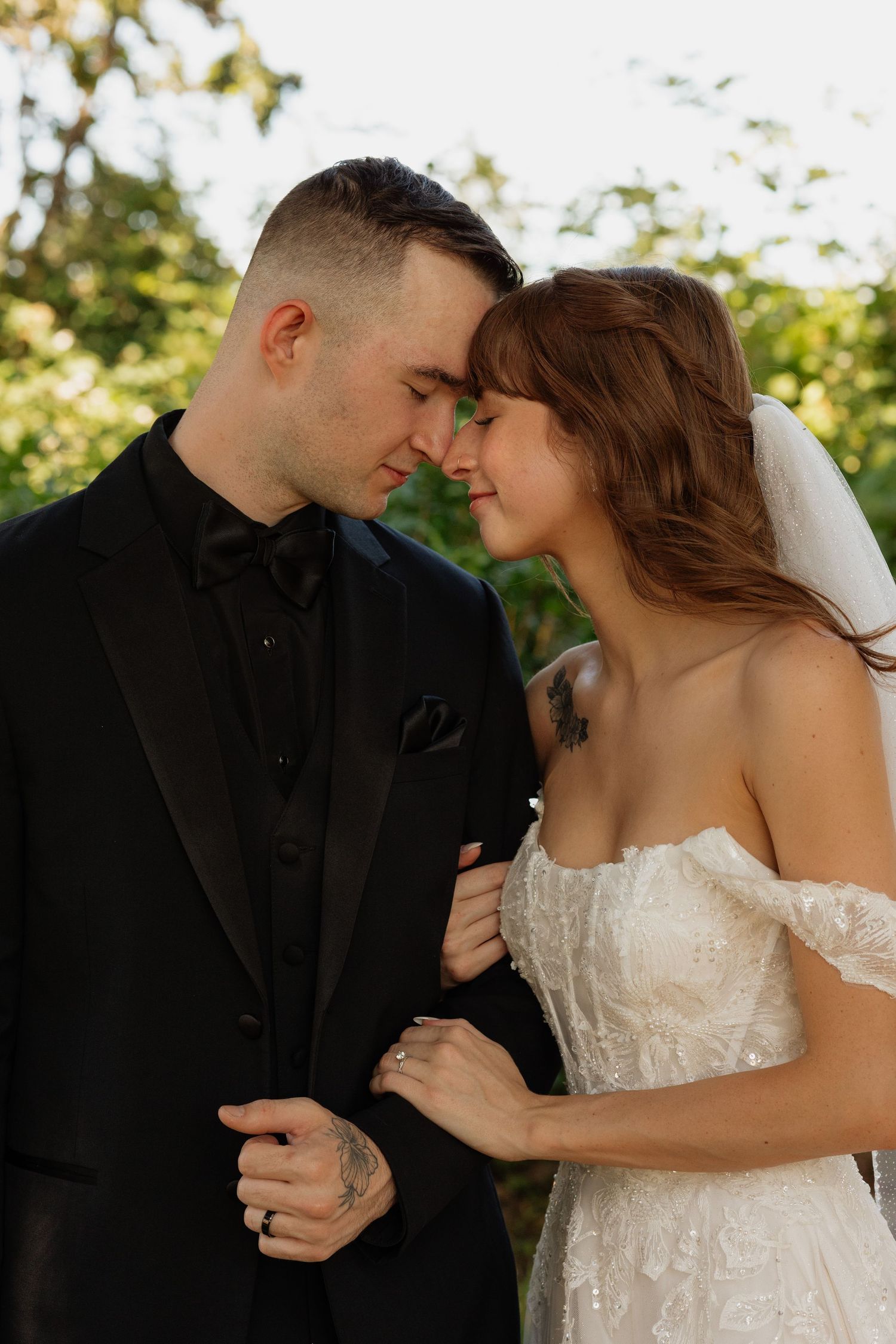 Bride and groom in black suit sharing an intimate forehead touch moment outdoors surrounded by green trees.