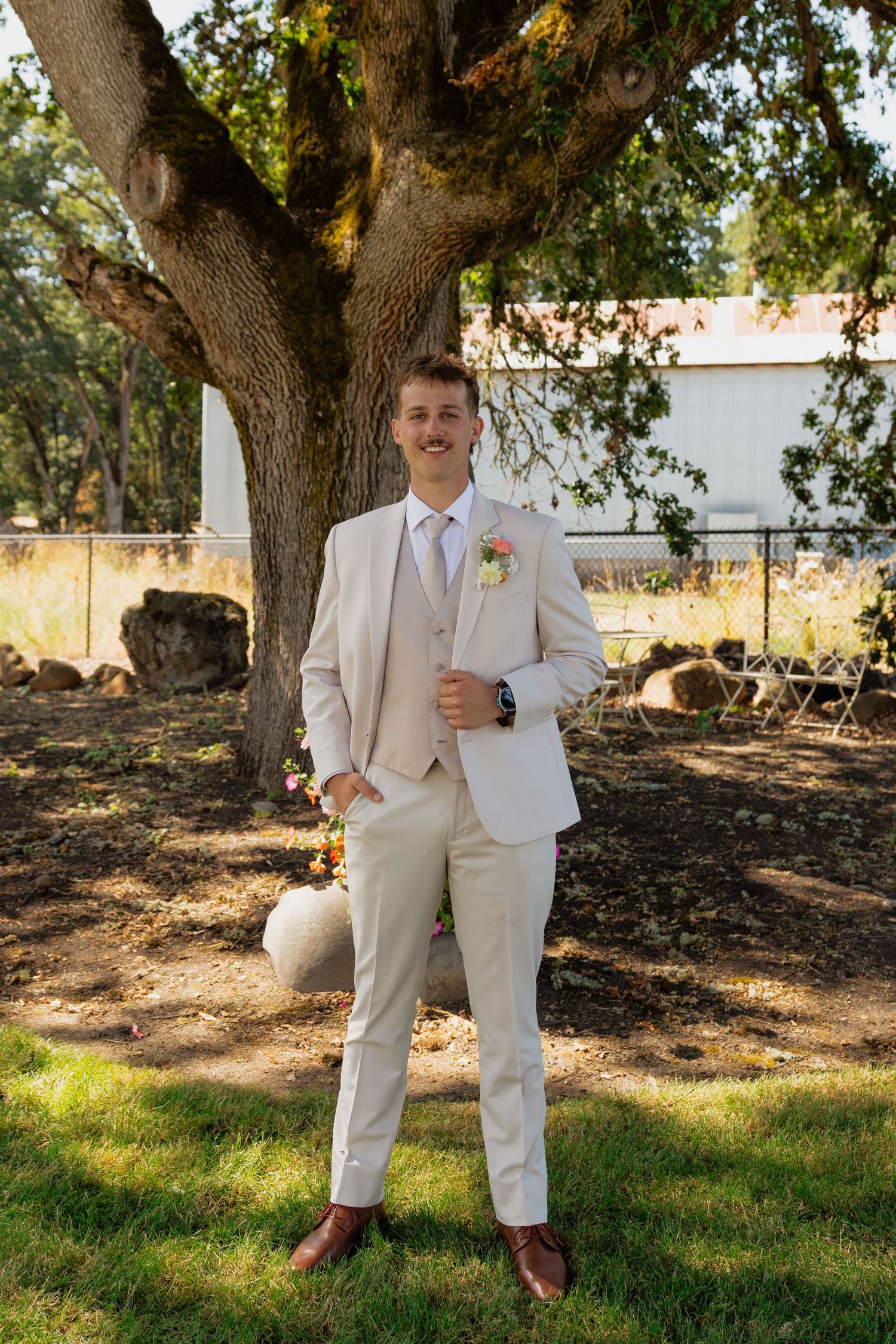 Groom in beige three-piece suit with boutonniere standing under large oak tree outdoors.