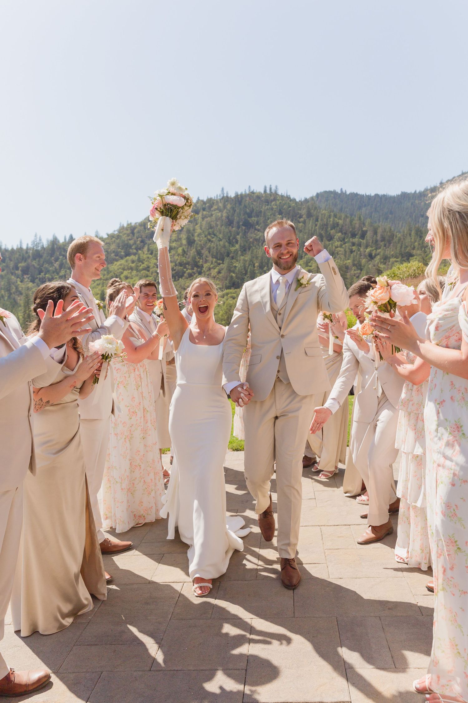Wedding party in neutral tones celebrates joyfully on a stone patio with mountains in the background.