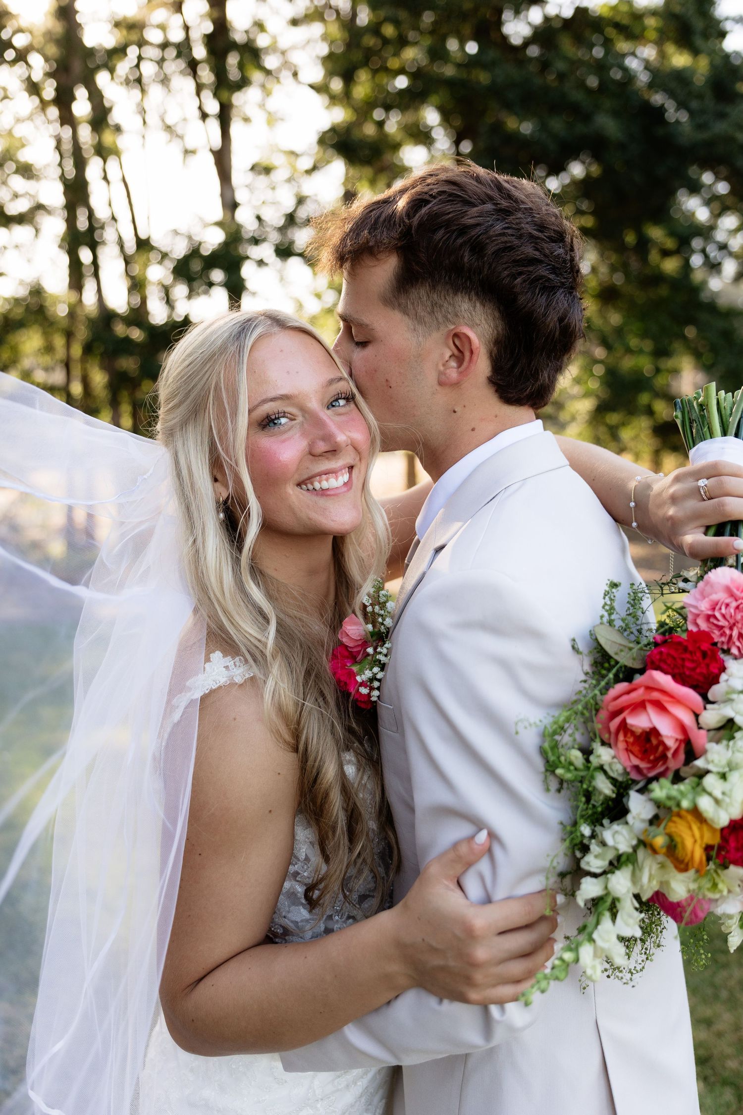 A smiling bride in a white gown holds a colorful bouquet as her groom in a white suit kisses her cheek outdoors.