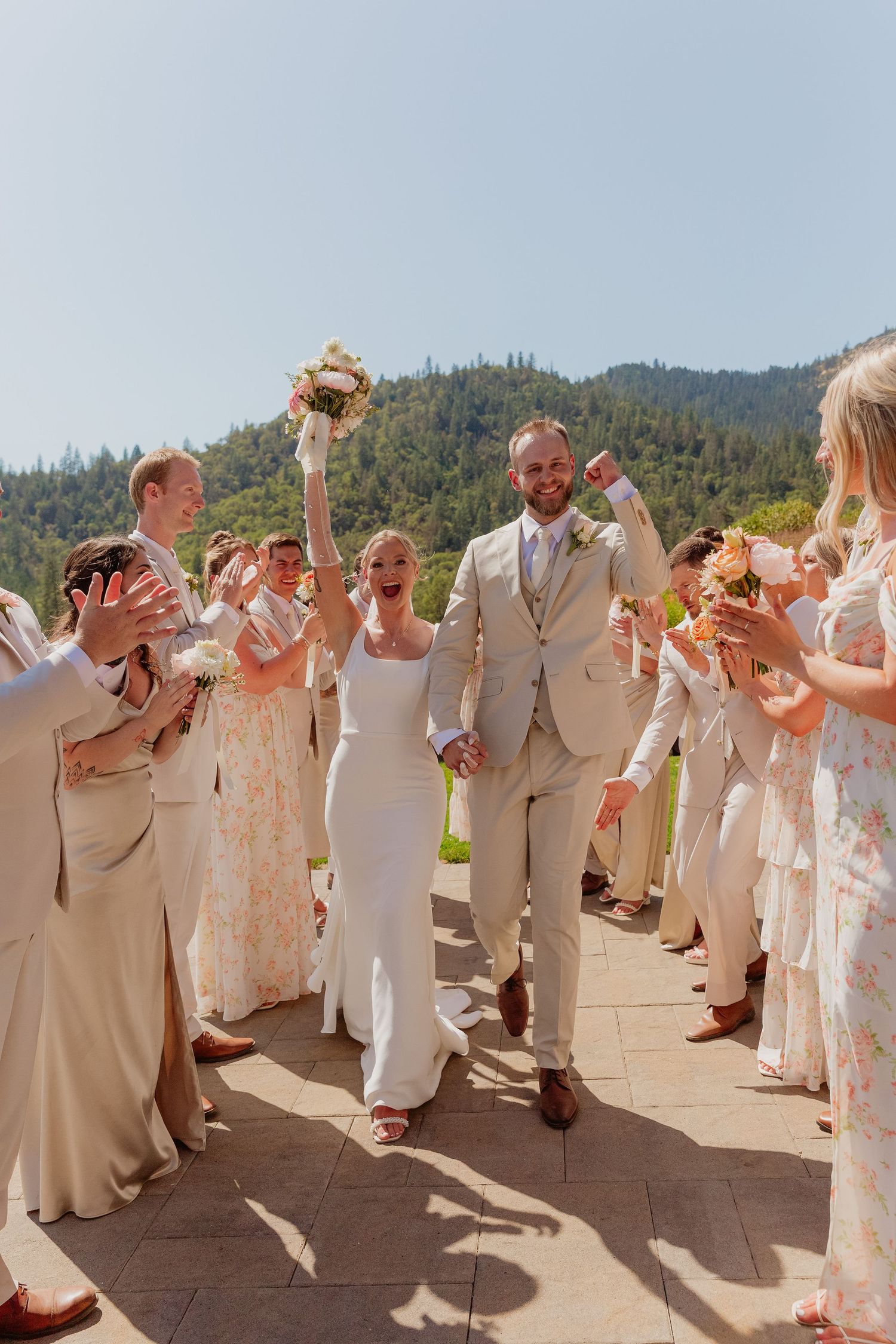 Joyful newlyweds walk through a celebratory aisle of guests at an outdoor wedding with lush green hills backdrop.