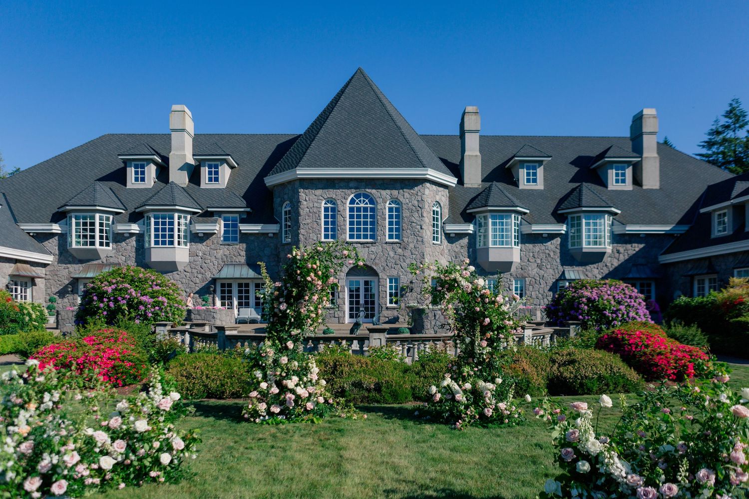 Luxury stone mansion with French-inspired architecture features dormer windows and vibrant flower gardens under blue sky.