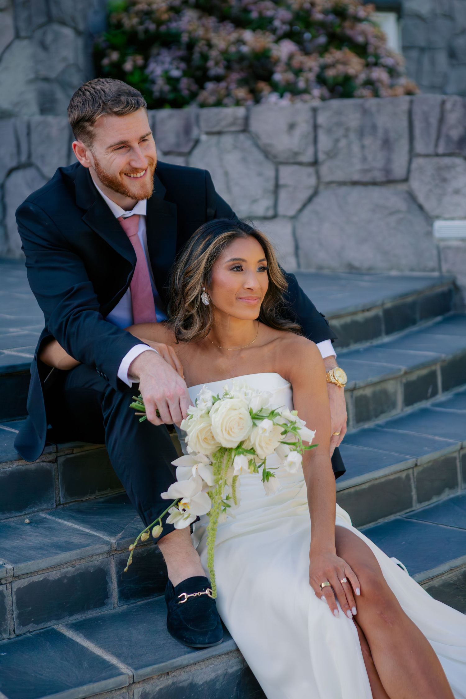 A couple sits together on stone steps, with the bride in white holding a bouquet of cream roses.
