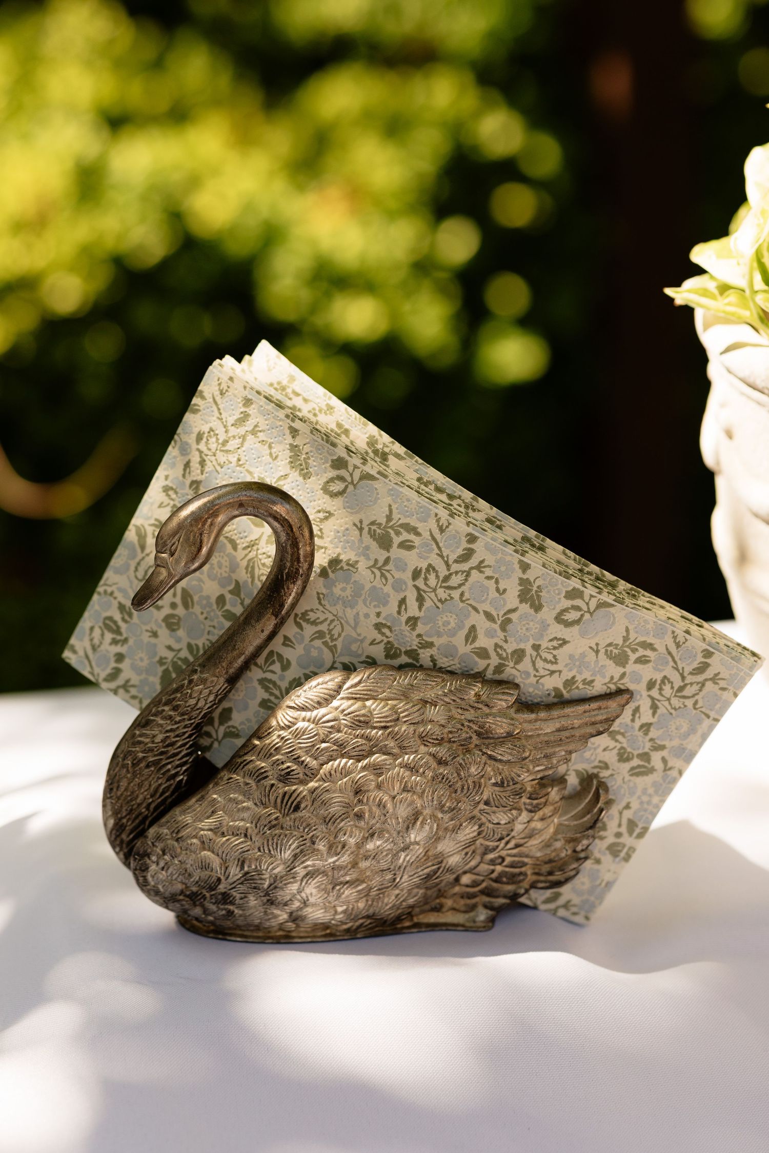 Antique brass swan napkin holder with floral patterned napkins on a white tablecloth at an outdoor event.