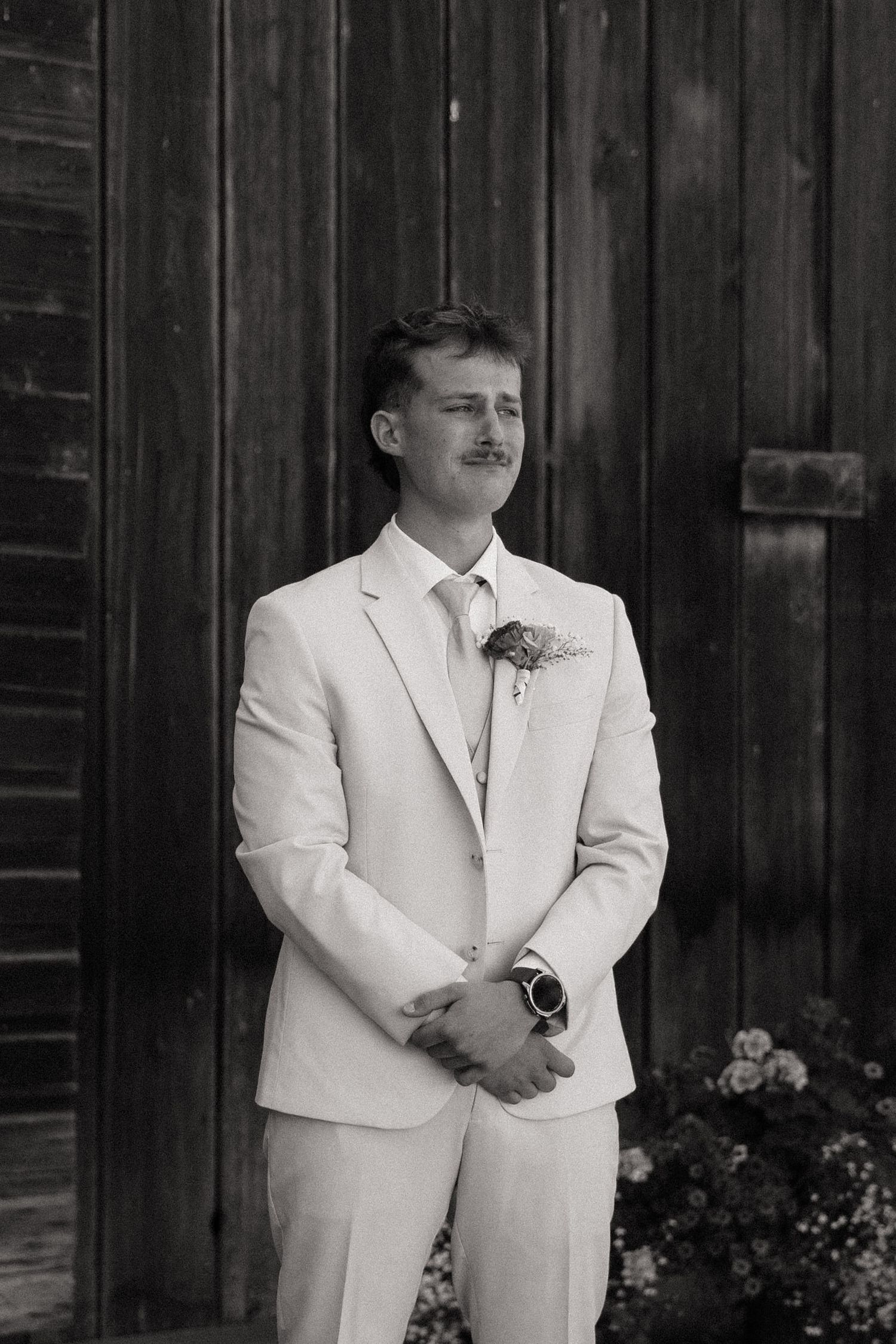 Groom in white suit with boutonniere stands confidently before rustic wooden barn doors in black and white wedding photo.