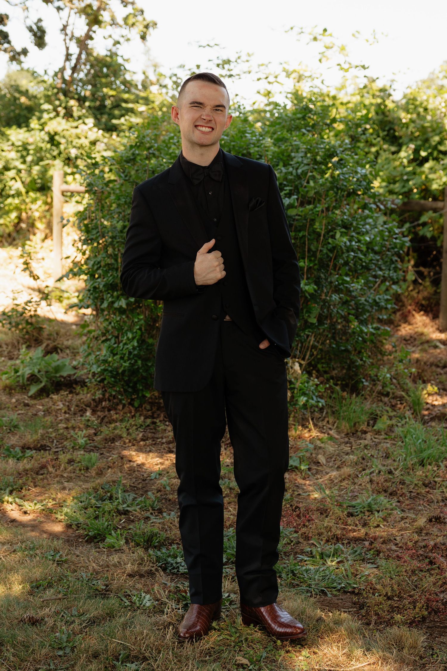 Groom in black suit smiling outdoors with lush green trees background.