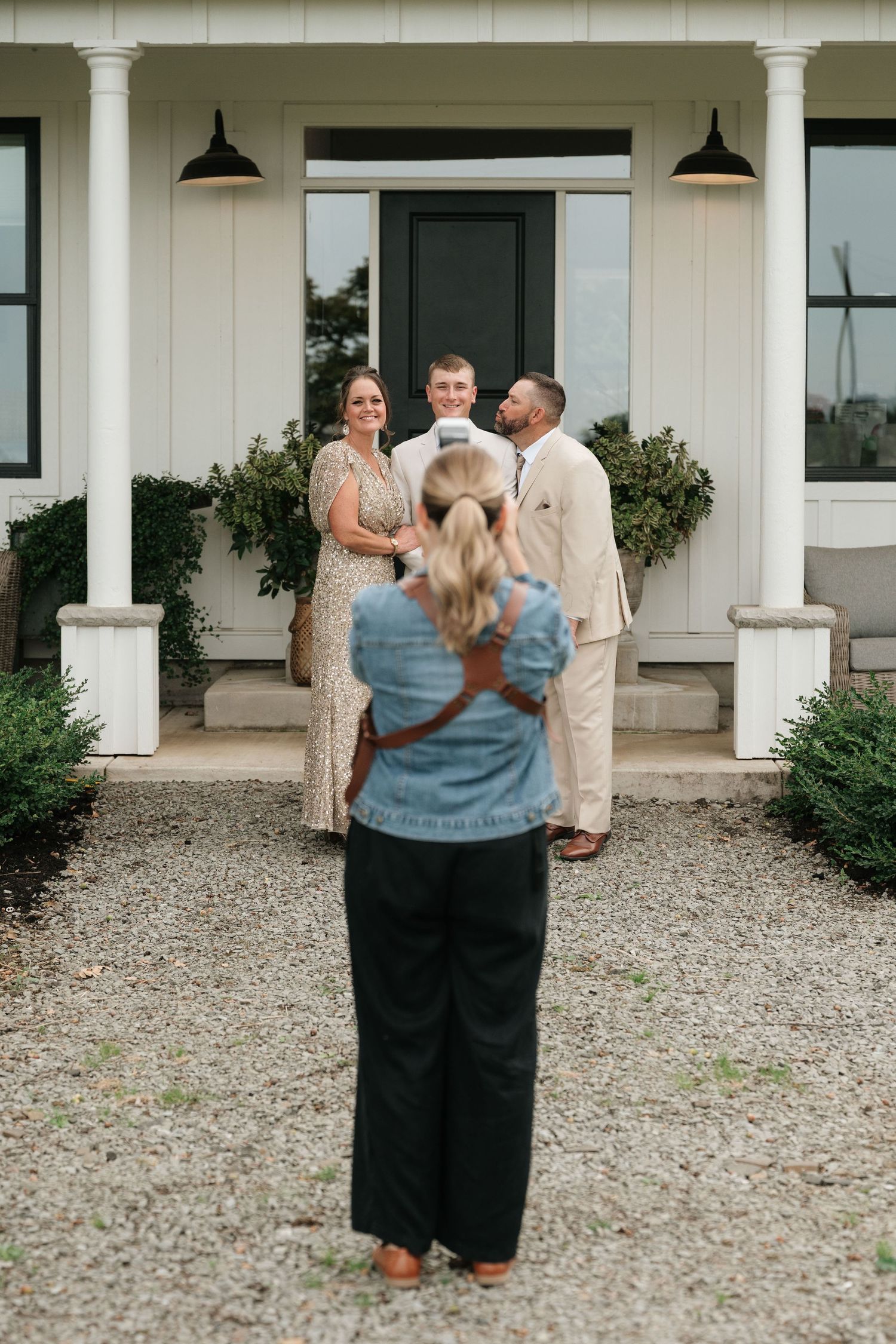 Photographer capturing a couple posing with a guest on a gravel path in front of a white farmhouse entrance.
