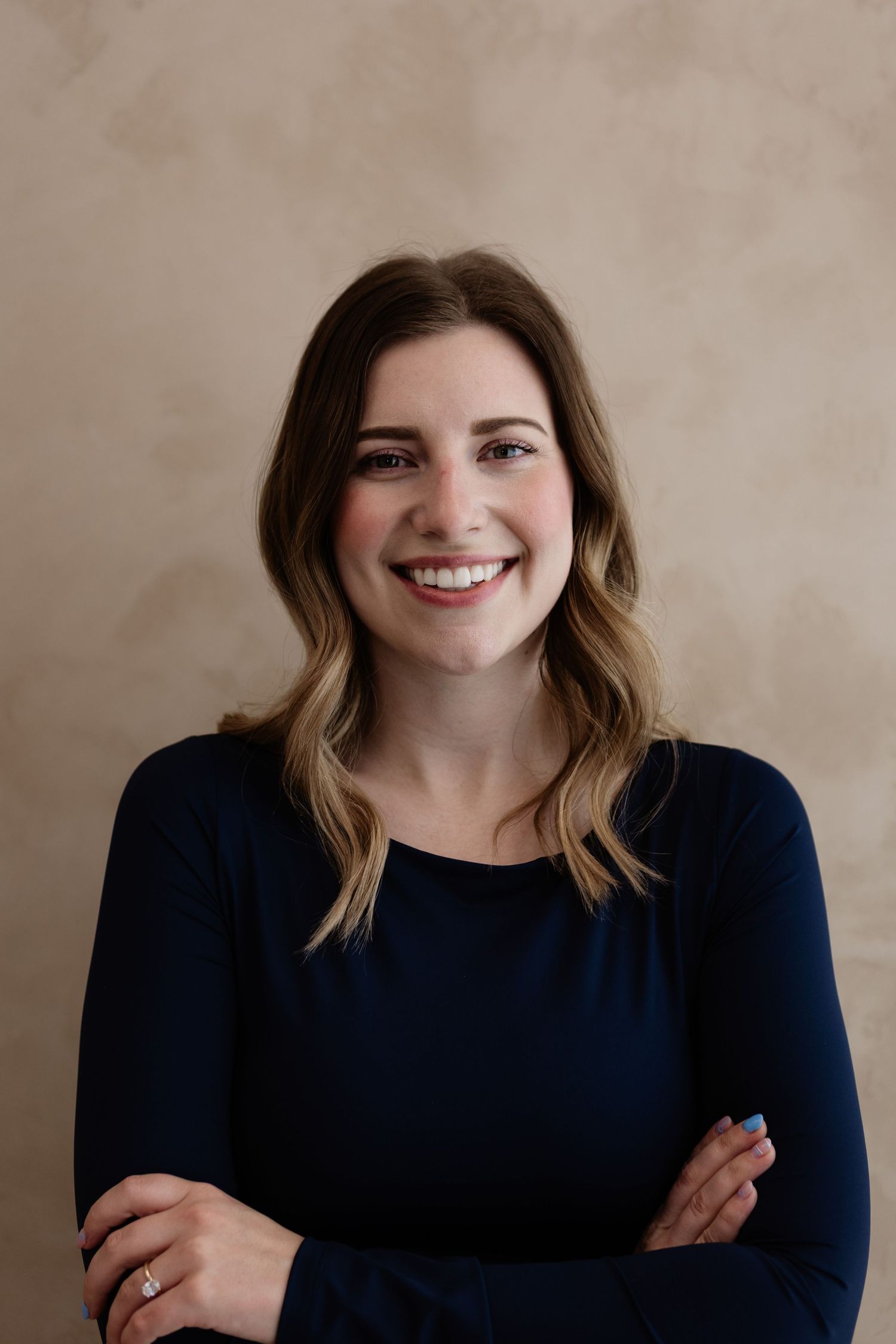 Smiling woman with brown hair in navy blue top, arms crossed, posing against a beige studio background.