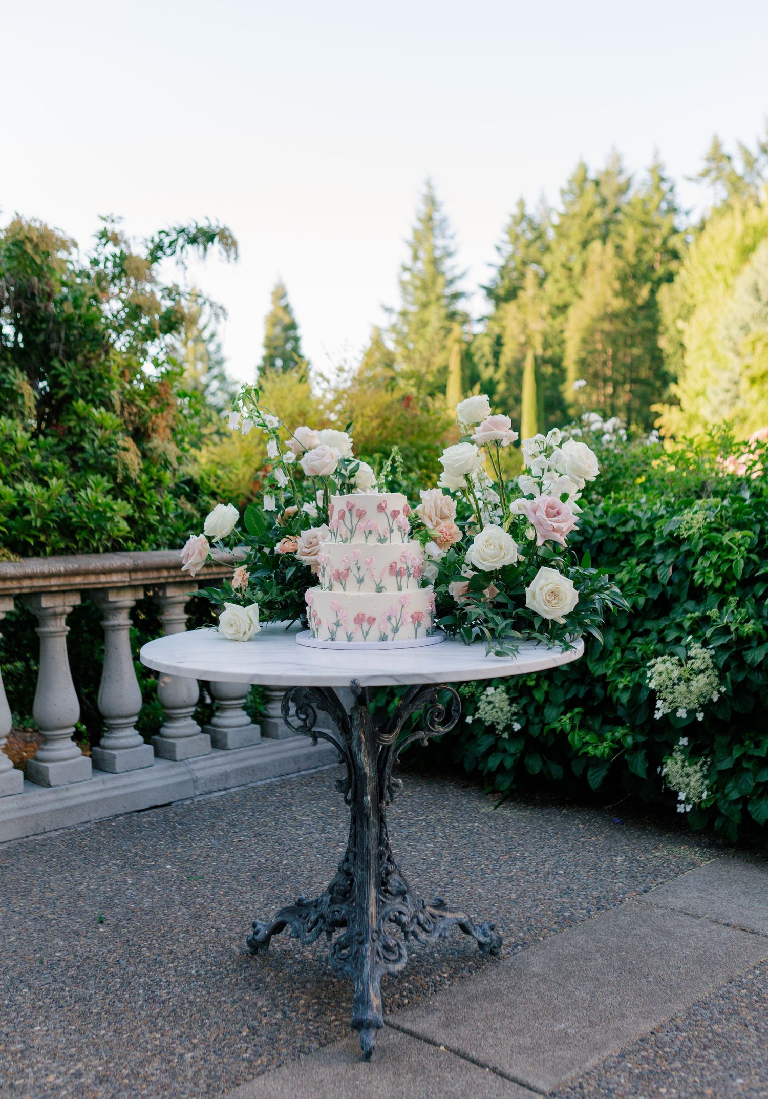 White floral centerpieces on ornate pedestals create an elegant outdoor garden display with pink and green accents.
