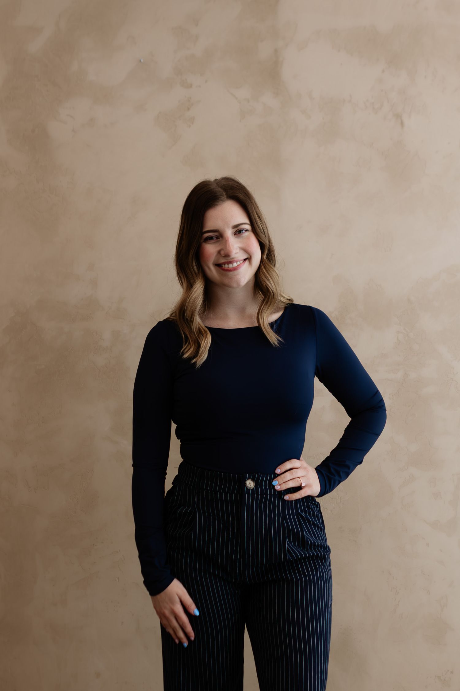 Smiling young woman in navy blue long-sleeve top and striped pants posing against a beige textured wall.
