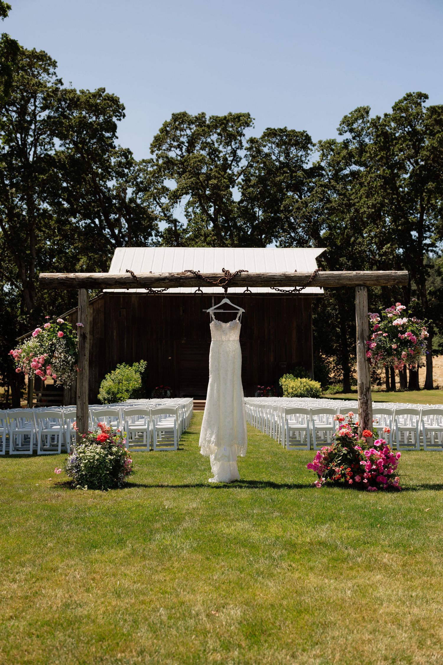 White wedding dress hanging on rustic wooden arch at outdoor ceremony venue with white chairs and pink floral arrangements.