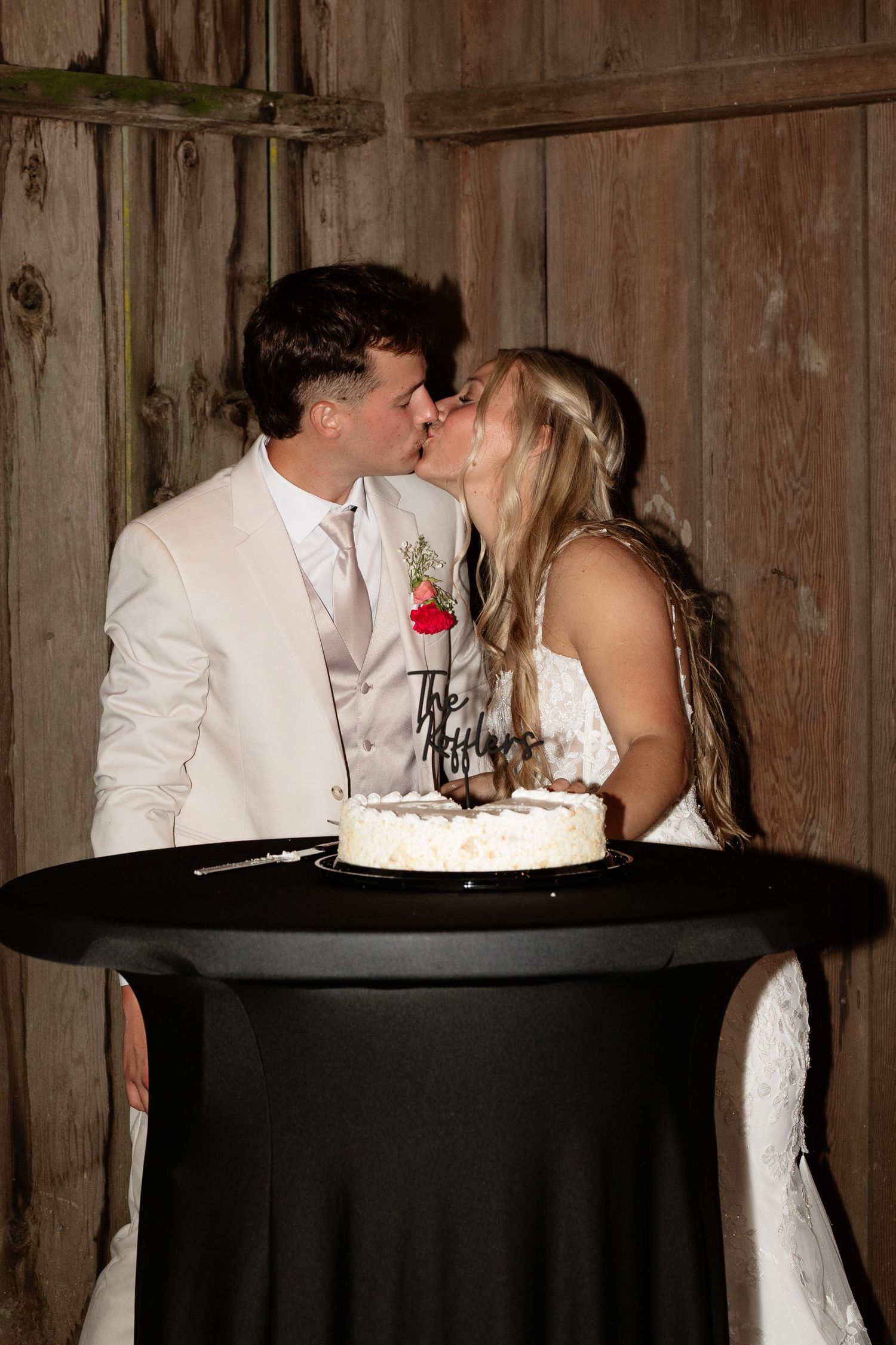 Newlyweds share a kiss over wedding cake at rustic barn reception with wooden backdrop.