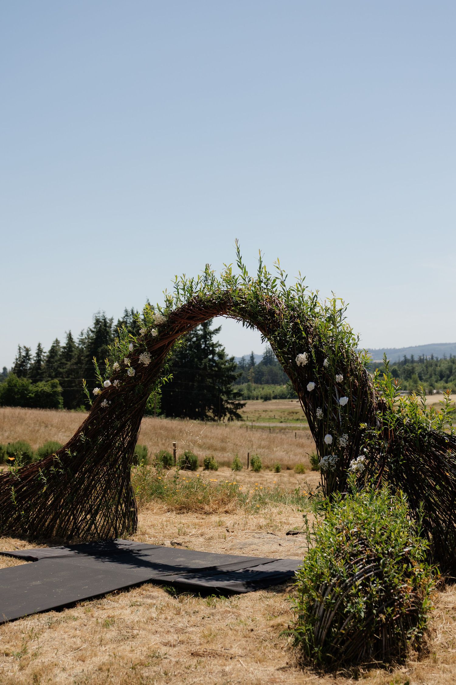Rustic woven branch wedding arch adorned with greenery and white flowers in an outdoor field setting.