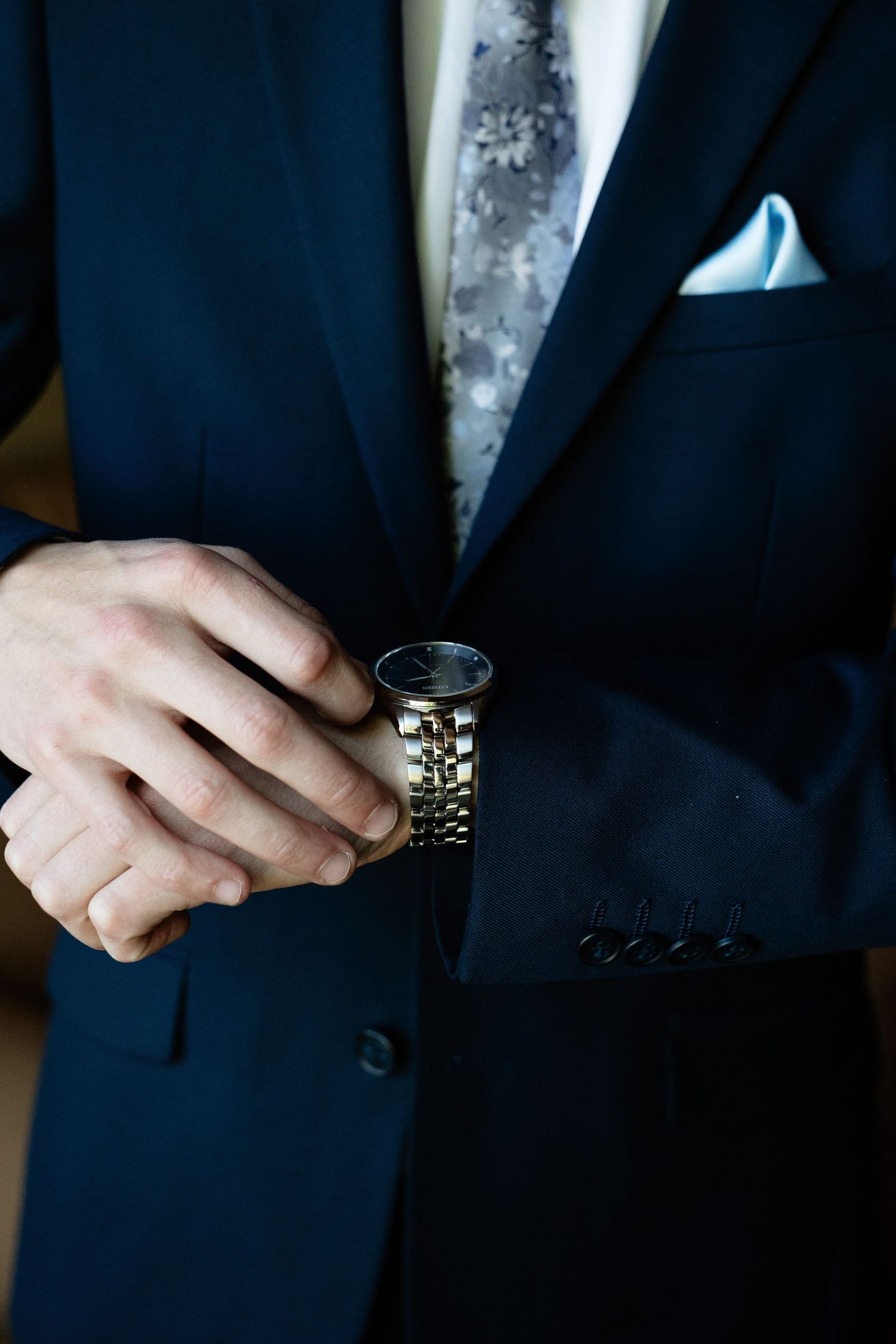 Groom in navy blue suit adjusting gold wristwatch, wearing floral tie and light blue pocket square.