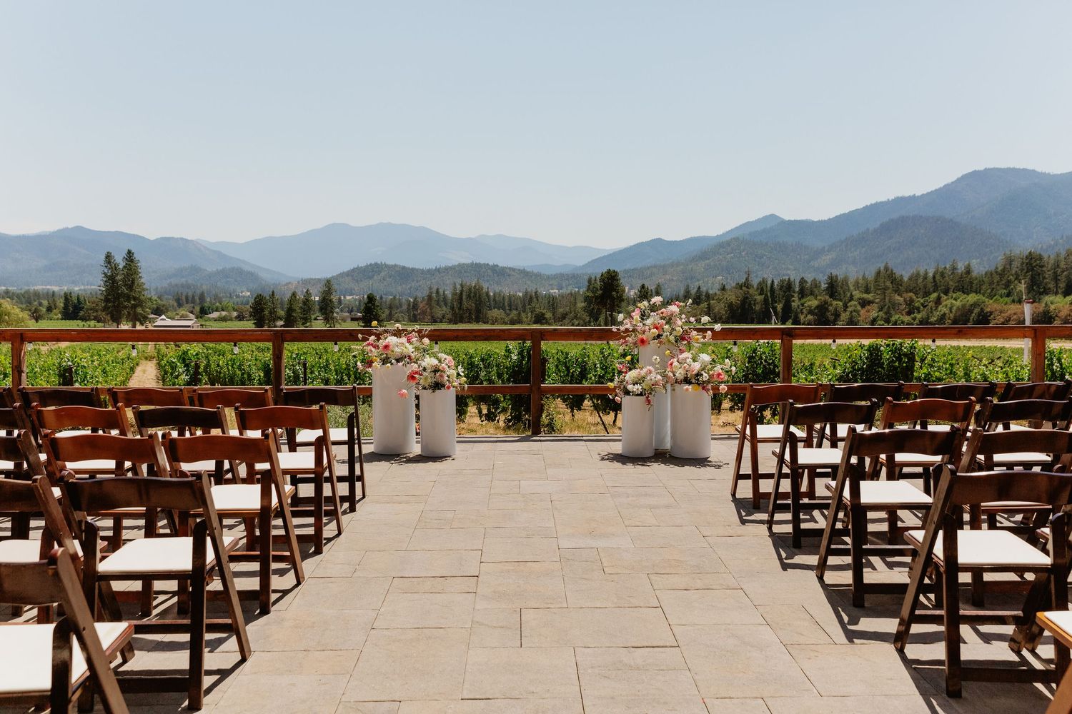 Outdoor vineyard wedding ceremony setup with wooden chairs, floral arrangements, and stunning mountain views under a clear blue sky.