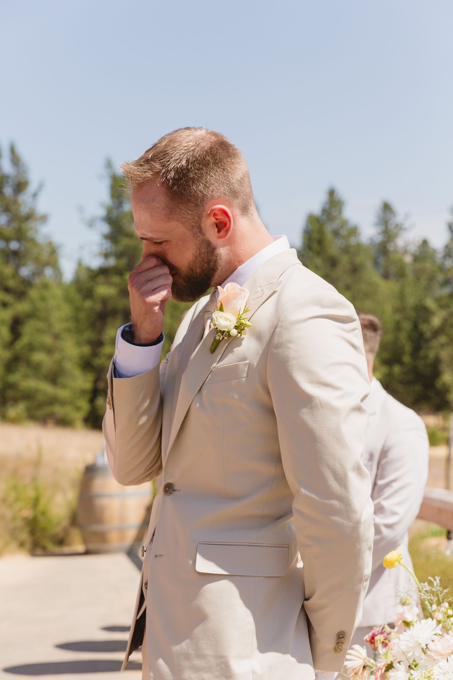 Groom in beige suit with yellow boutonniere shows emotional reaction during outdoor wedding ceremony.