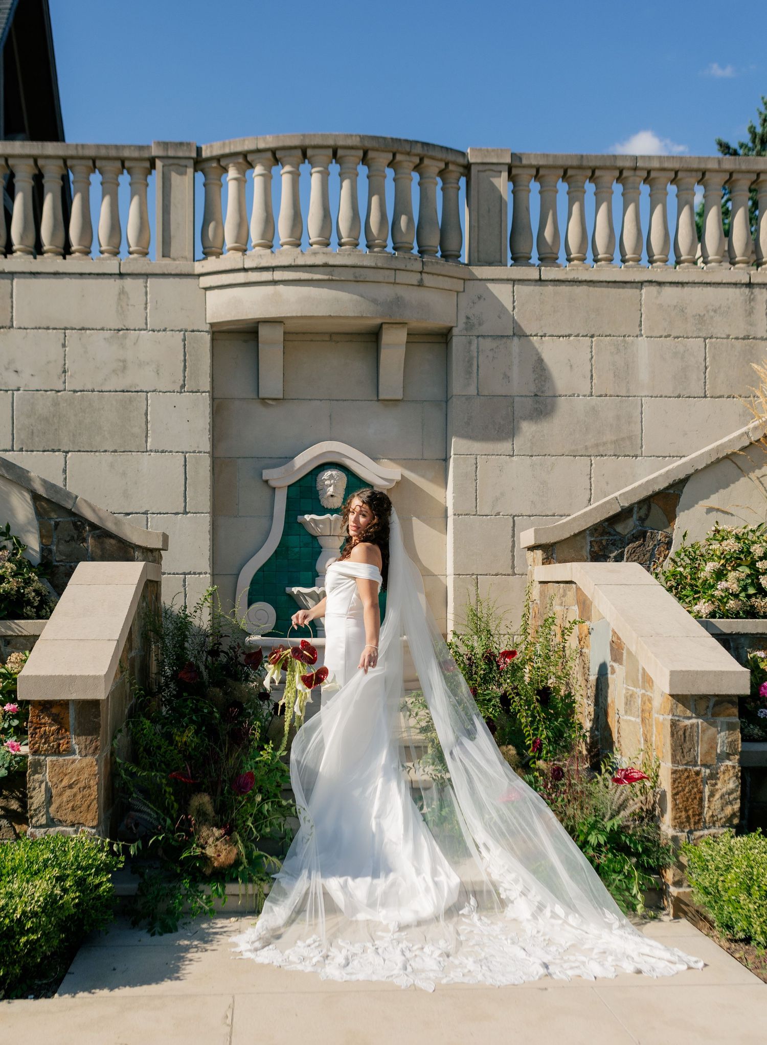 A wedding dress with a flowing veil photographed in sequence against a stone wall and garden landscape.