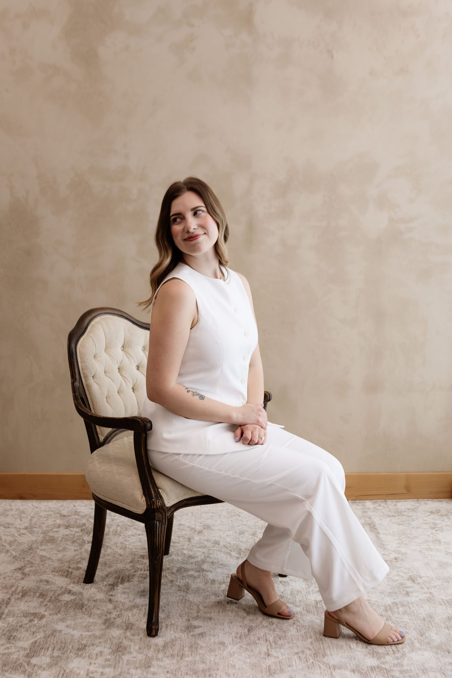 Woman in white outfit sitting gracefully on a vintage tufted chair against a beige textured wall.
