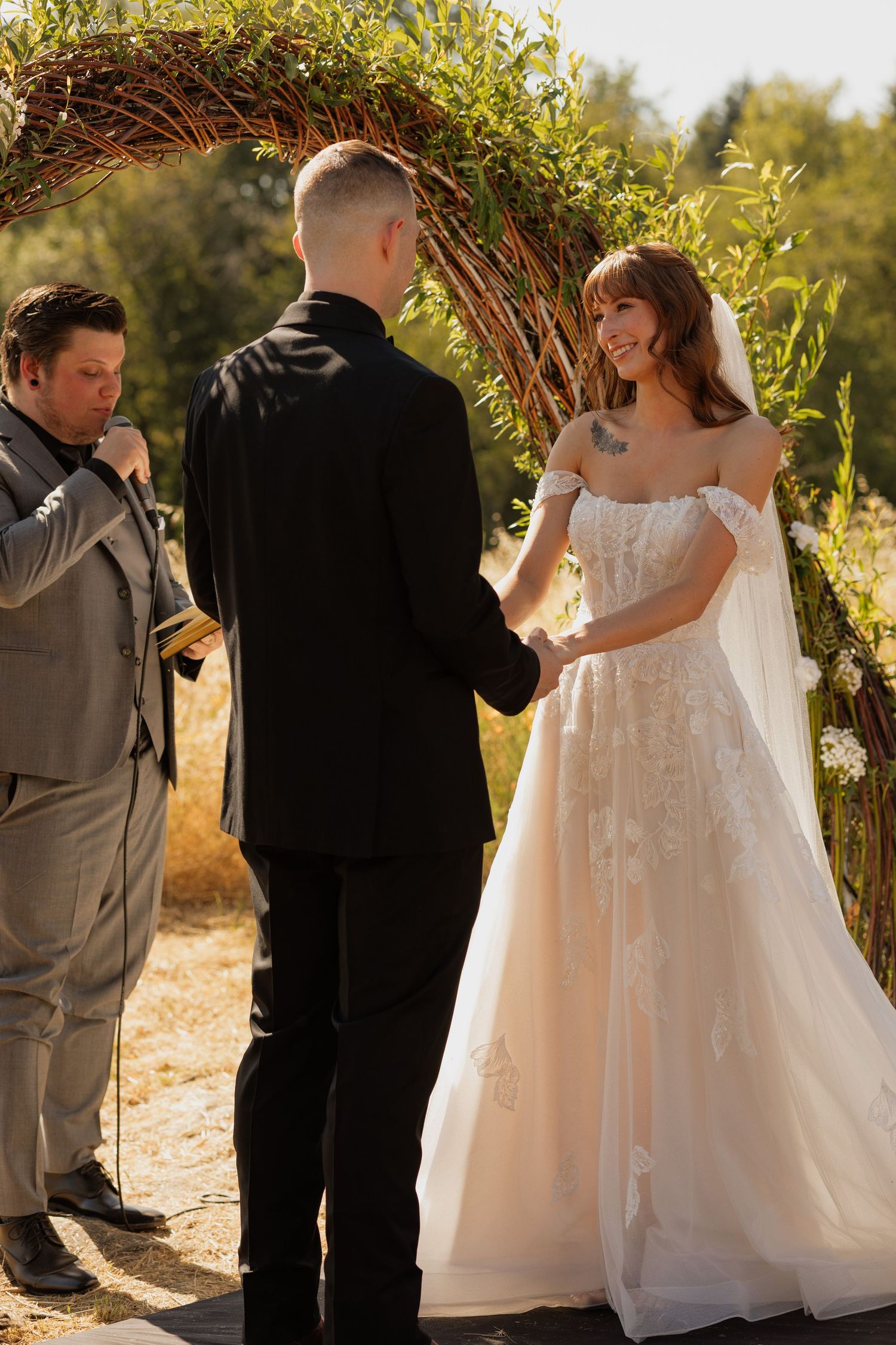 Bride and groom exchange vows under a greenery arch outdoors, officiant reads from book at sunny wedding ceremony.