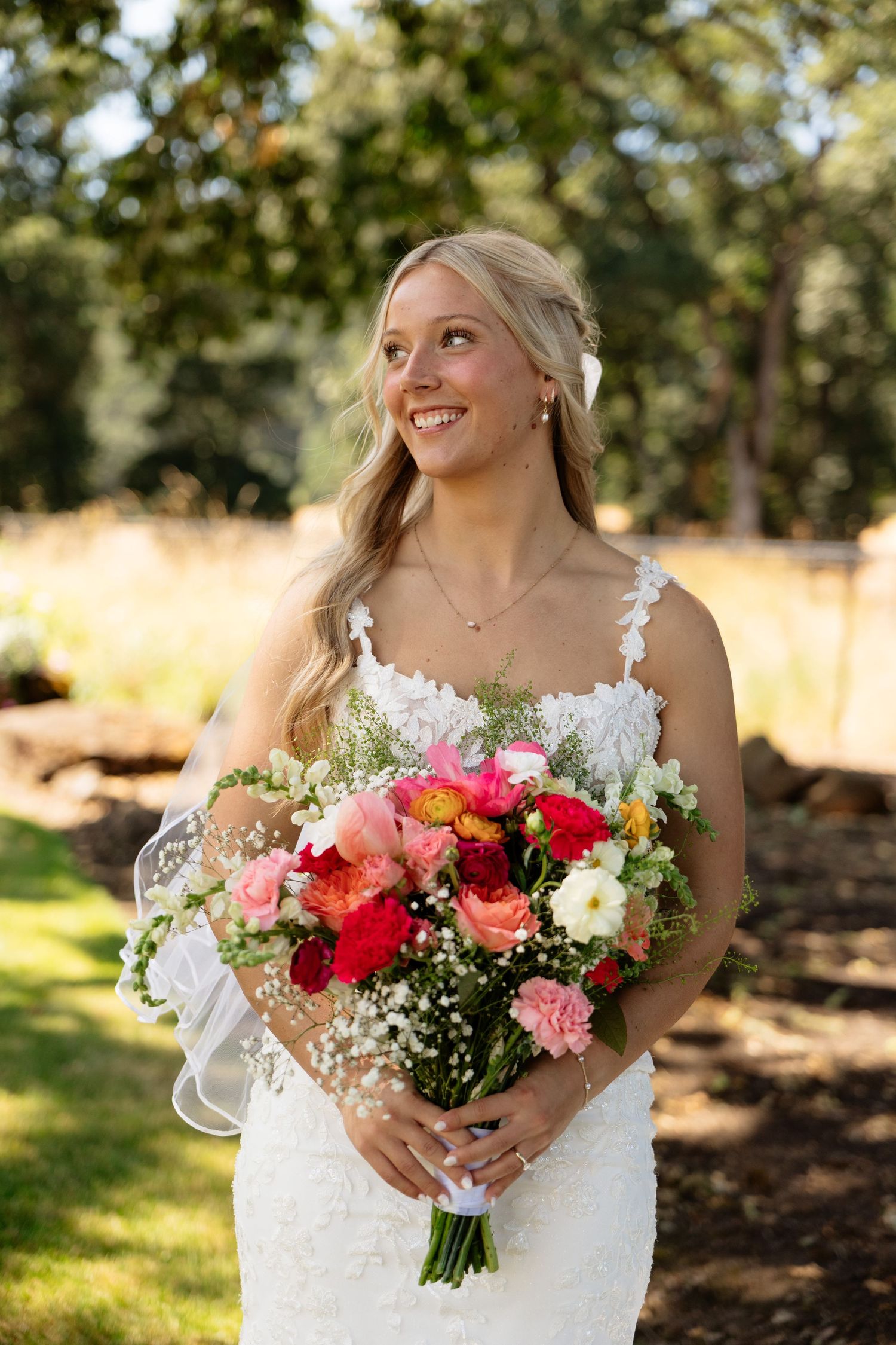 Smiling bride in lace wedding dress holding colorful bouquet of pink and red flowers outdoors.