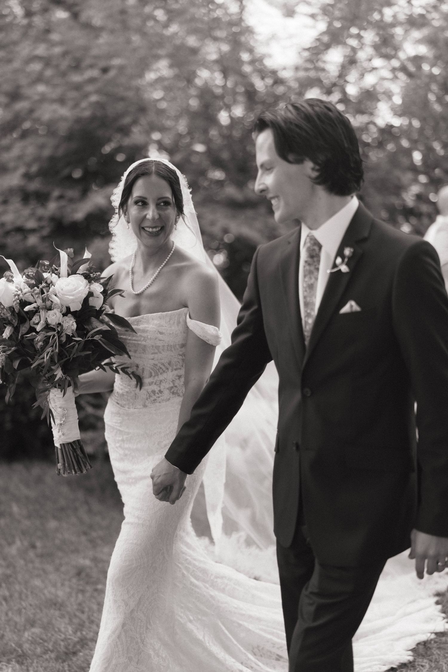 Black and white photo of smiling bride in lace gown with veil and bouquet walking with groom outdoors.