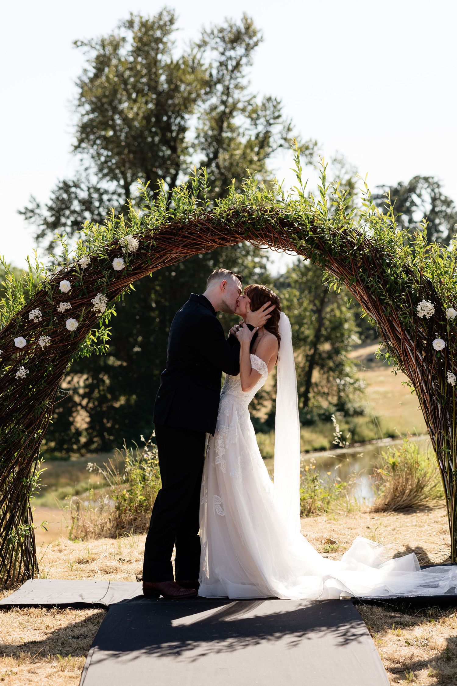Bride and groom share first kiss under a rustic floral arch at an outdoor wedding ceremony.