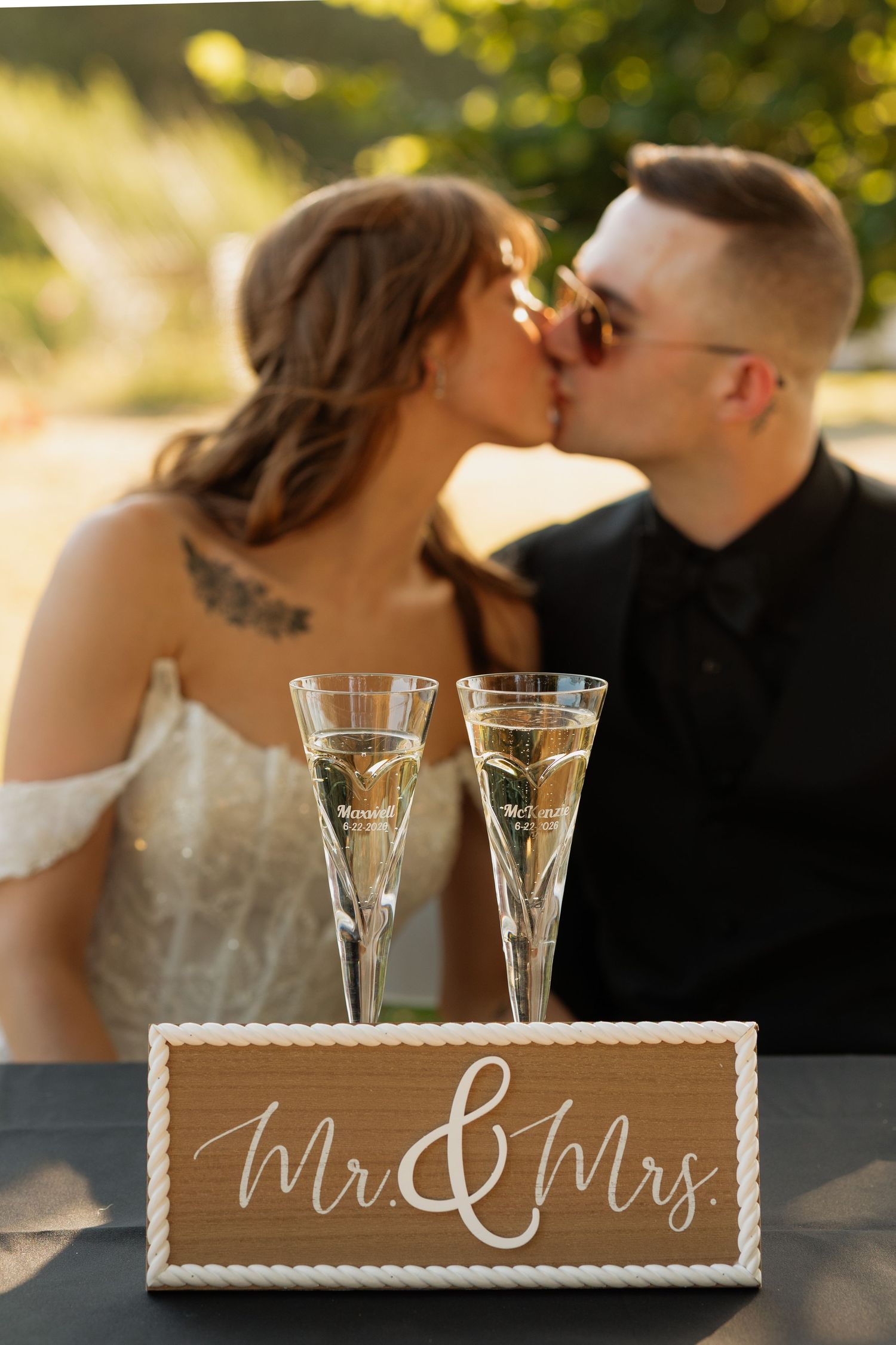 Bride and groom kissing behind Mr. & Mrs. sign with champagne flutes at outdoor wedding reception.