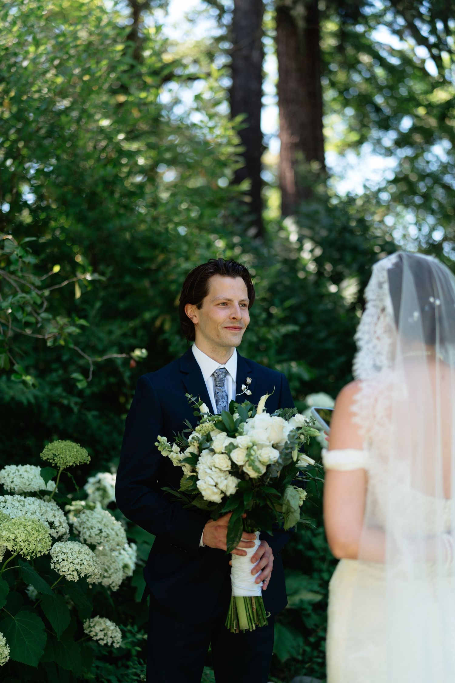 Groom in navy suit smiles at bride during outdoor first look moment surrounded by white hydrangeas and lush greenery.
