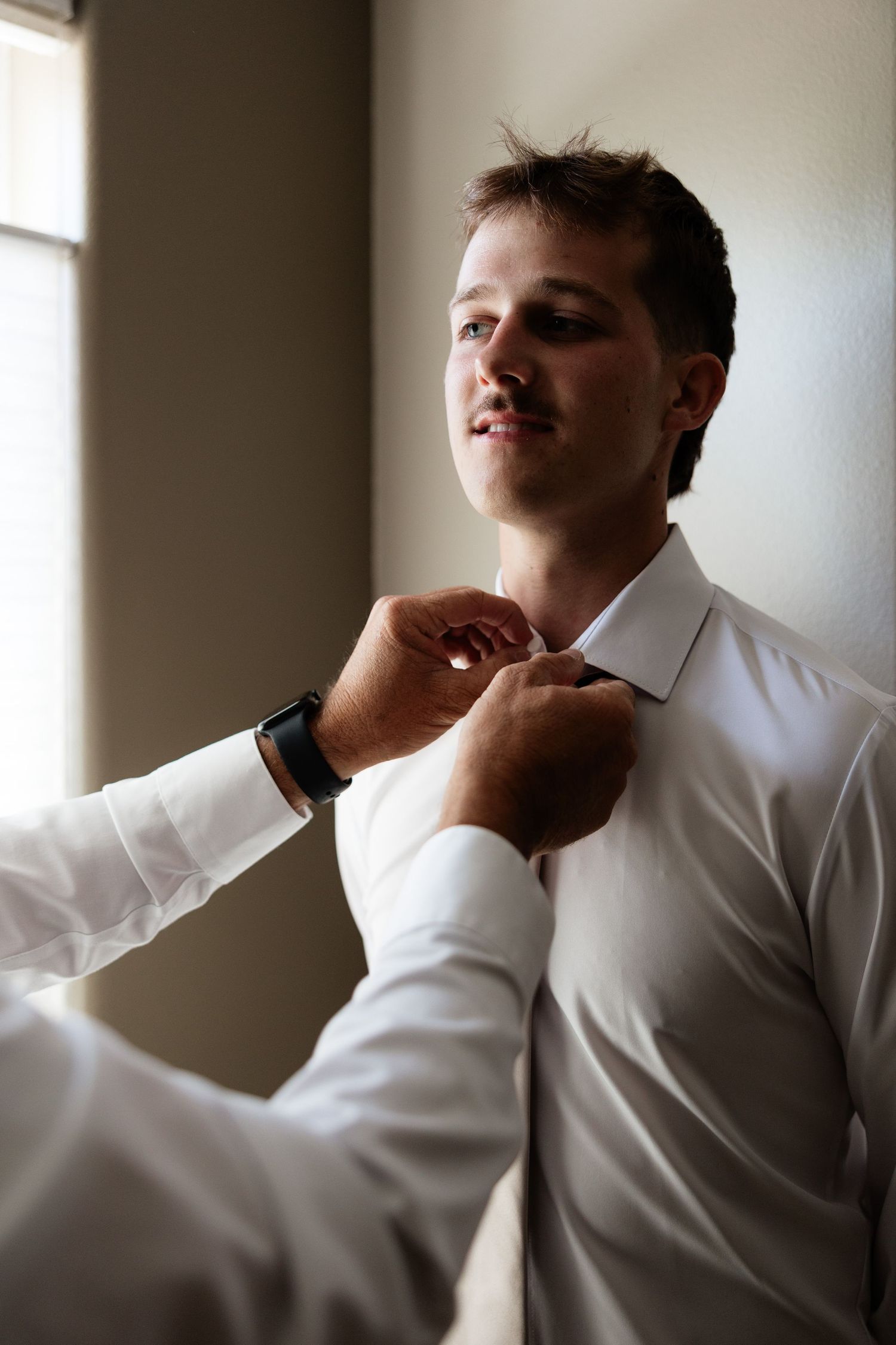 Groomsman helping groom button white dress shirt collar near window before wedding ceremony.