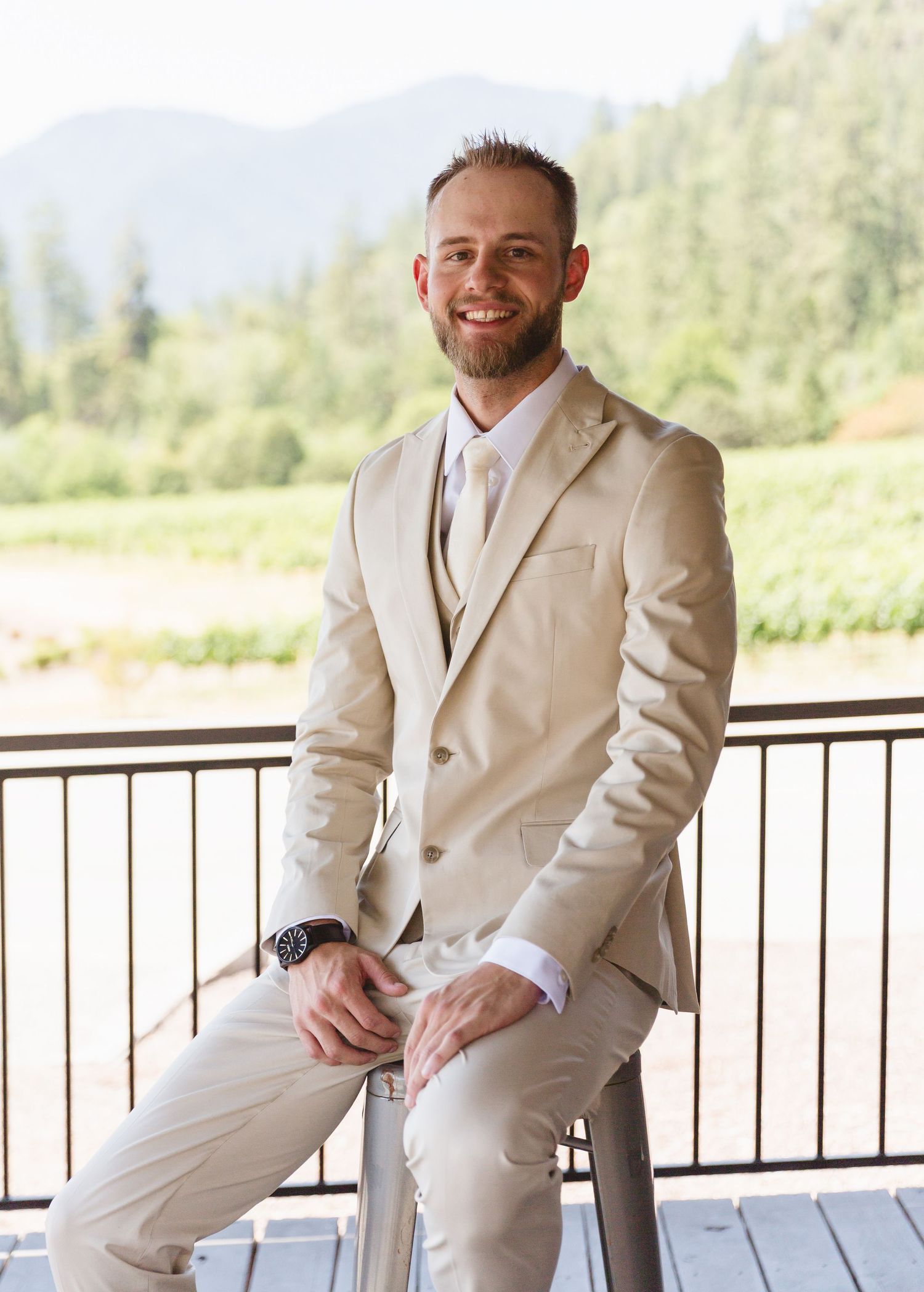 Groom wearing a beige three-piece suit smiles while sitting on a railing with mountain scenery in the background.