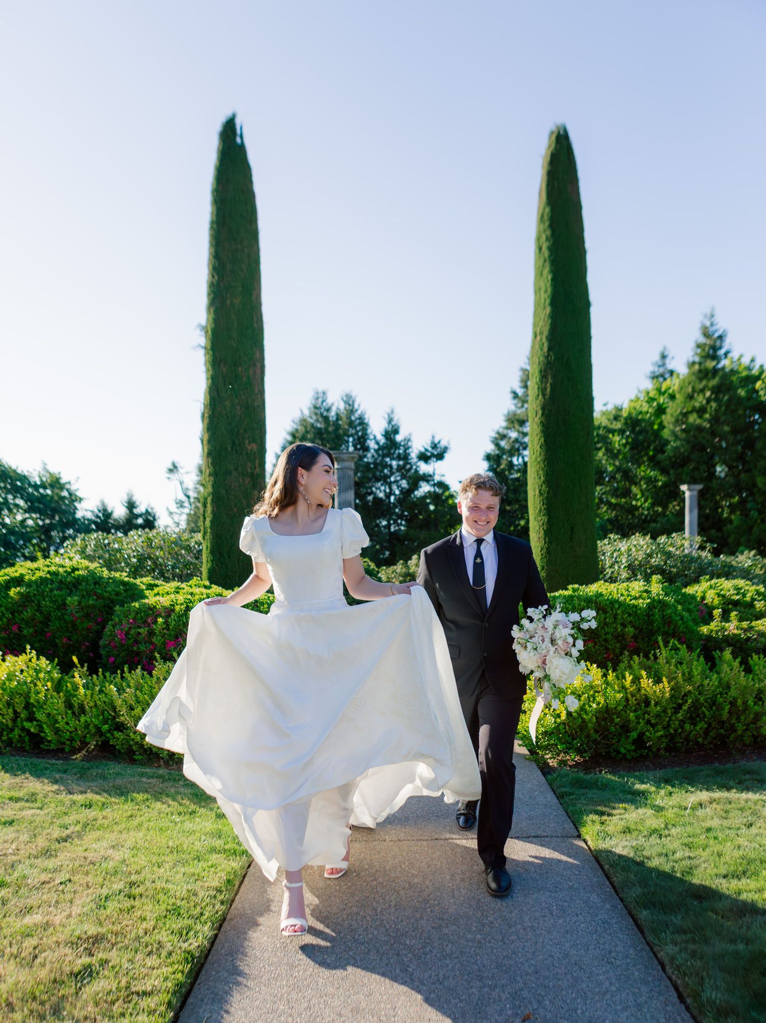 A couple walks along a garden path between tall cypress trees, with the bride lifting her flowing white dress.