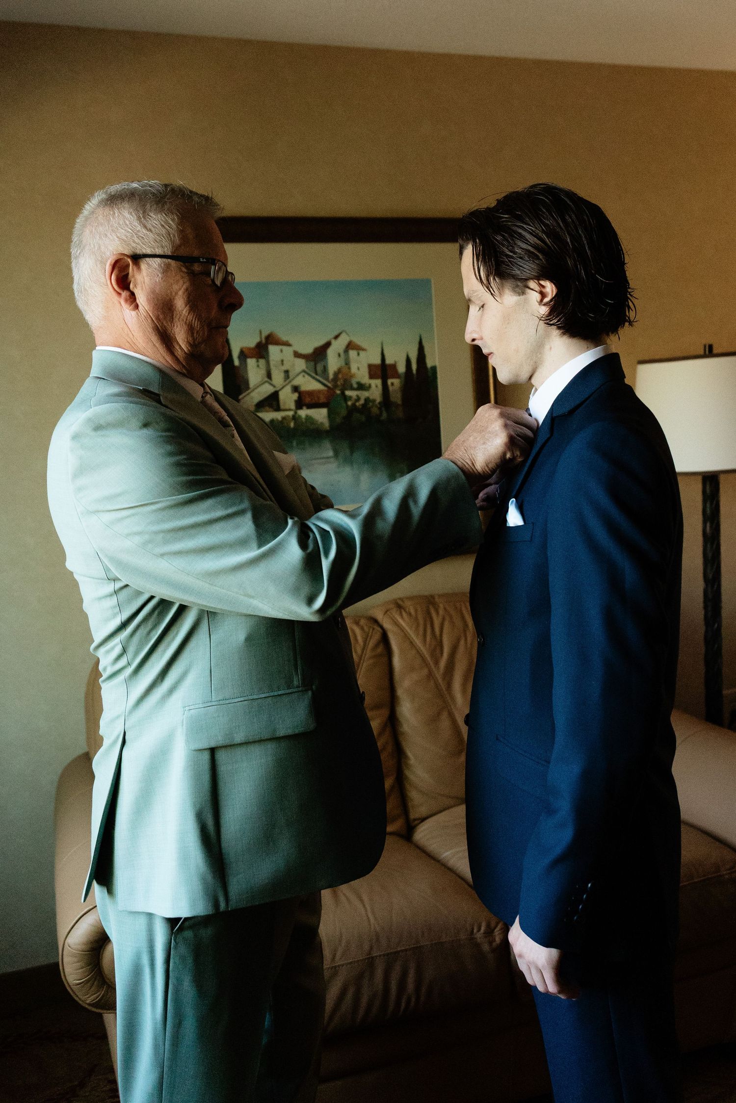 Father helping groom adjust navy suit tie in hotel room before wedding ceremony.