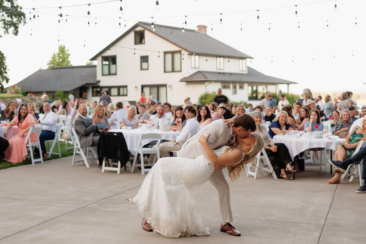 Bride and groom share romantic dip kiss on outdoor dance floor at elegant wedding reception with guests seated at tables.
