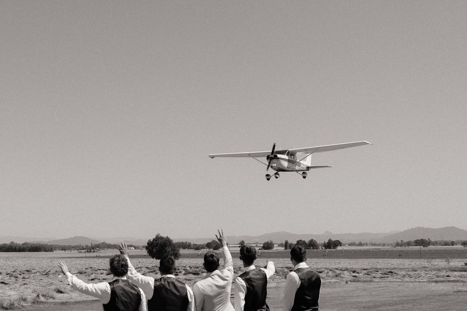 Group of people waving goodbye to a small propeller plane taking off from a rural airstrip in black and white.