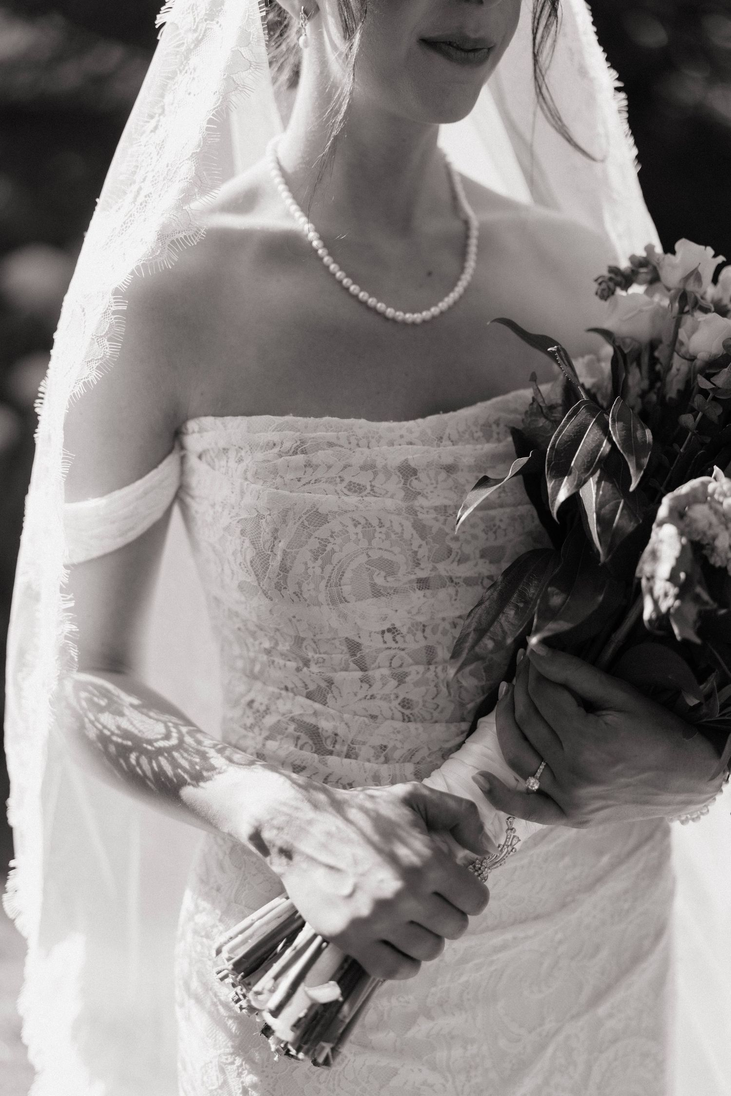 Black and white bridal portrait showing strapless lace gown, pearl necklace, veil, tattooed arm, and bouquet.