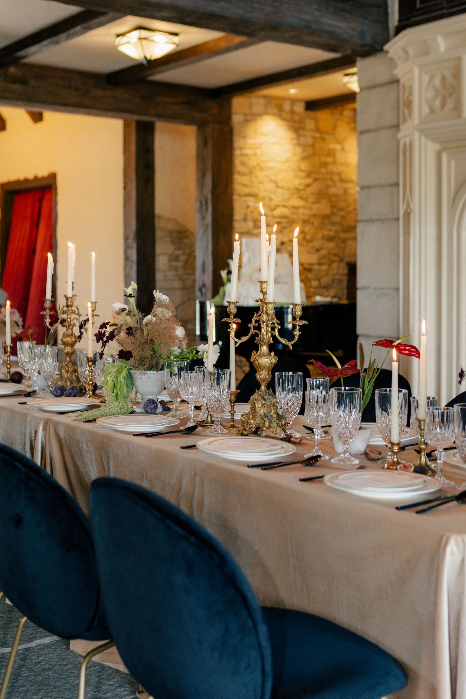Elegant dining table setting with crystal candelabras, white linens and navy blue velvet chairs in a stone-walled room.