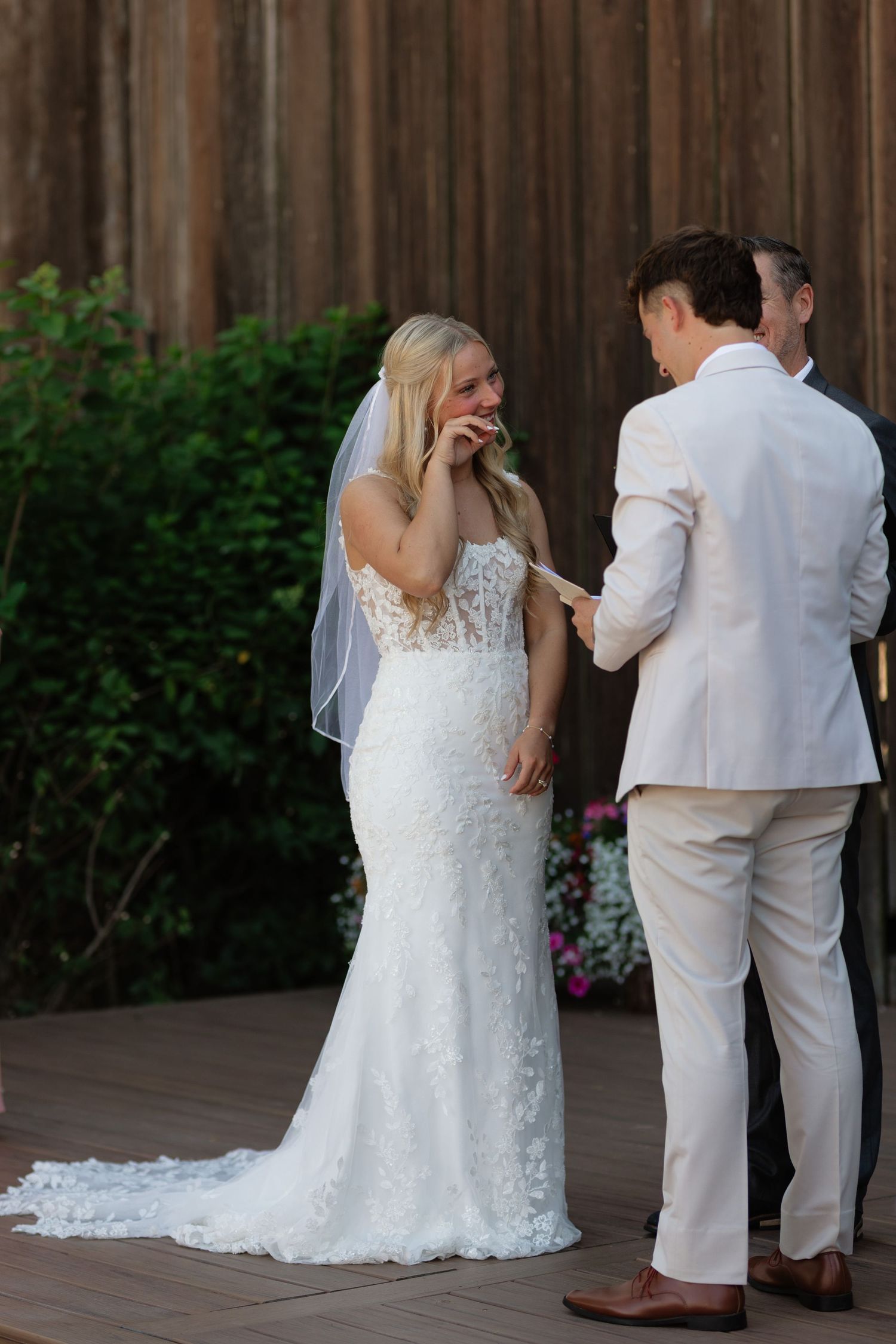 Emotional bride wipes tears during outdoor wedding ceremony while groom reads vows at rustic venue.
