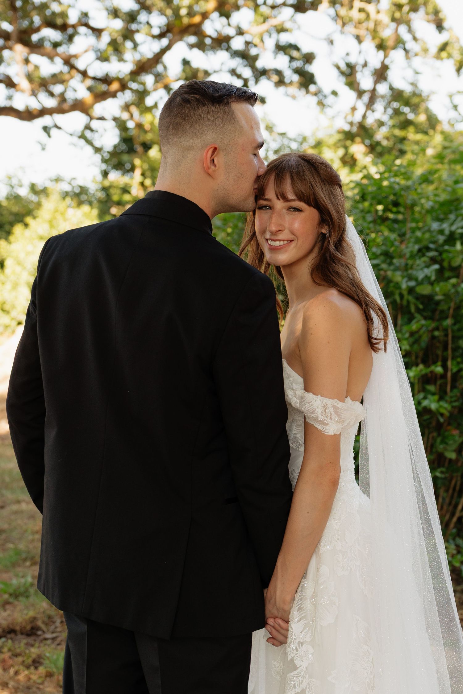 Smiling bride in off-shoulder lace gown with veil, groom in black suit kissing her cheek outdoors.