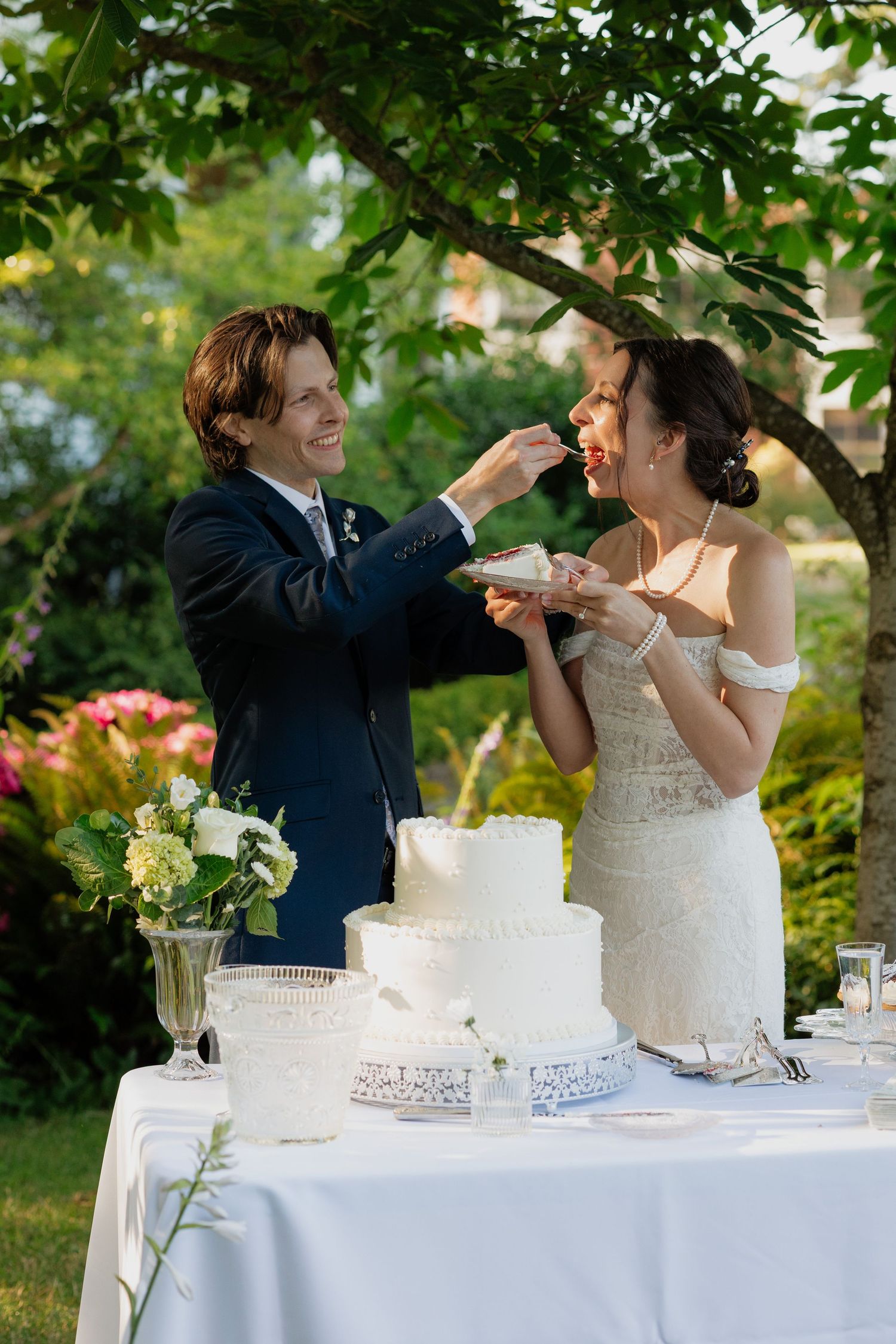 Smiling groom feeds bride wedding cake slice at outdoor garden reception table.