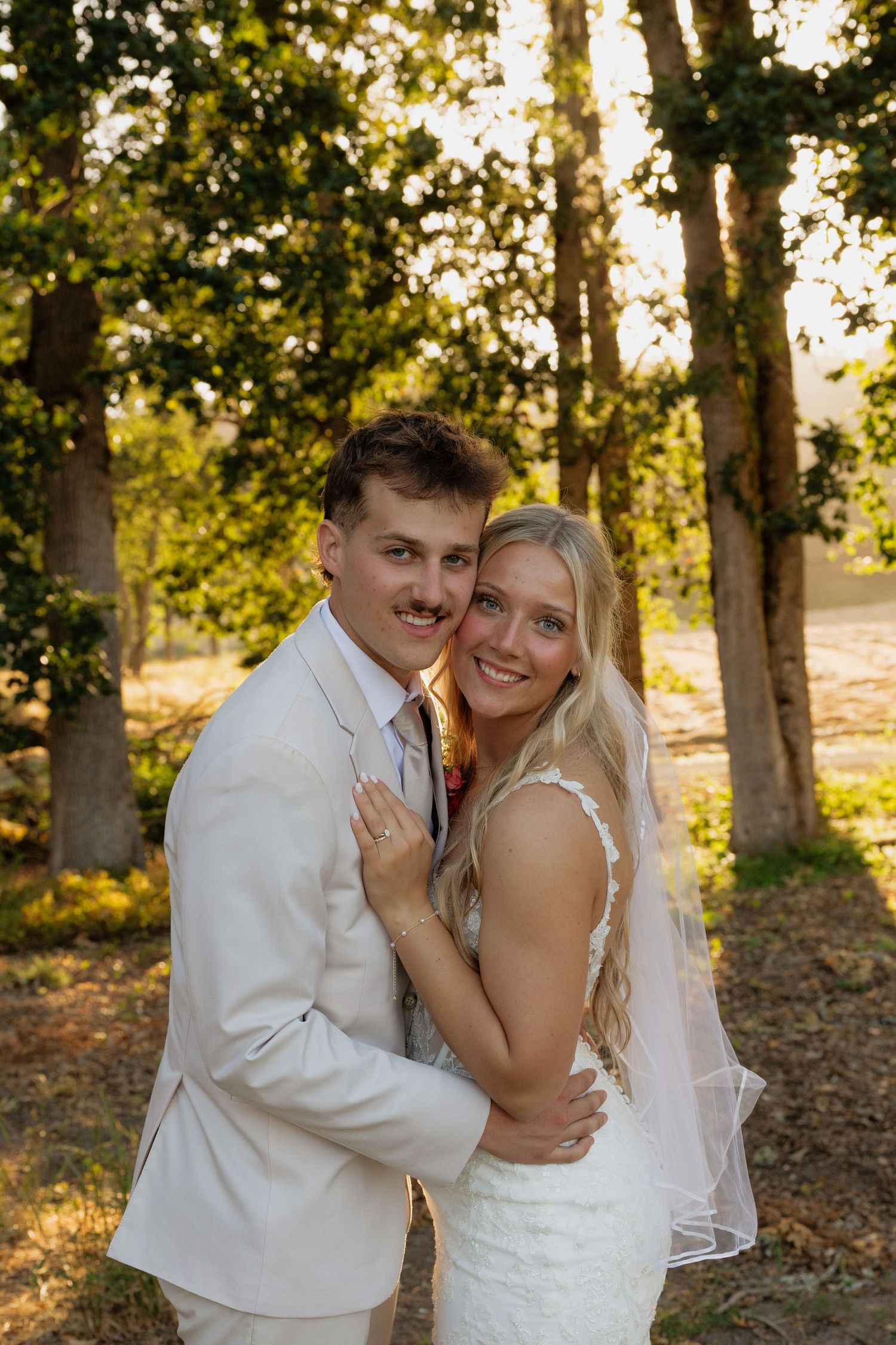 Smiling newlyweds embrace warmly among sunlit trees in romantic golden hour wedding portrait.