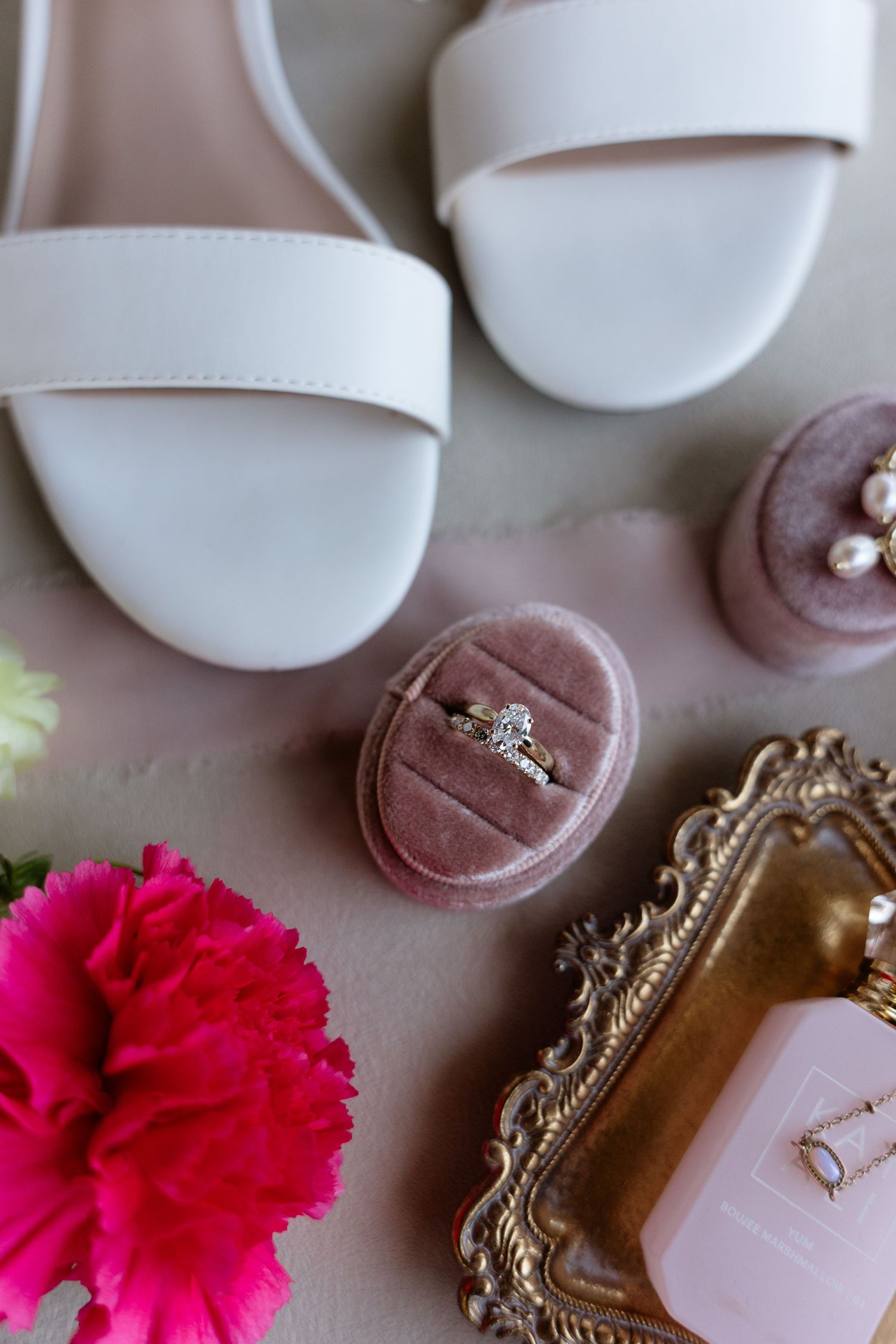 Bridal flat lay with white heels, diamond ring in pink velvet box, pearl earrings, and pink carnation.