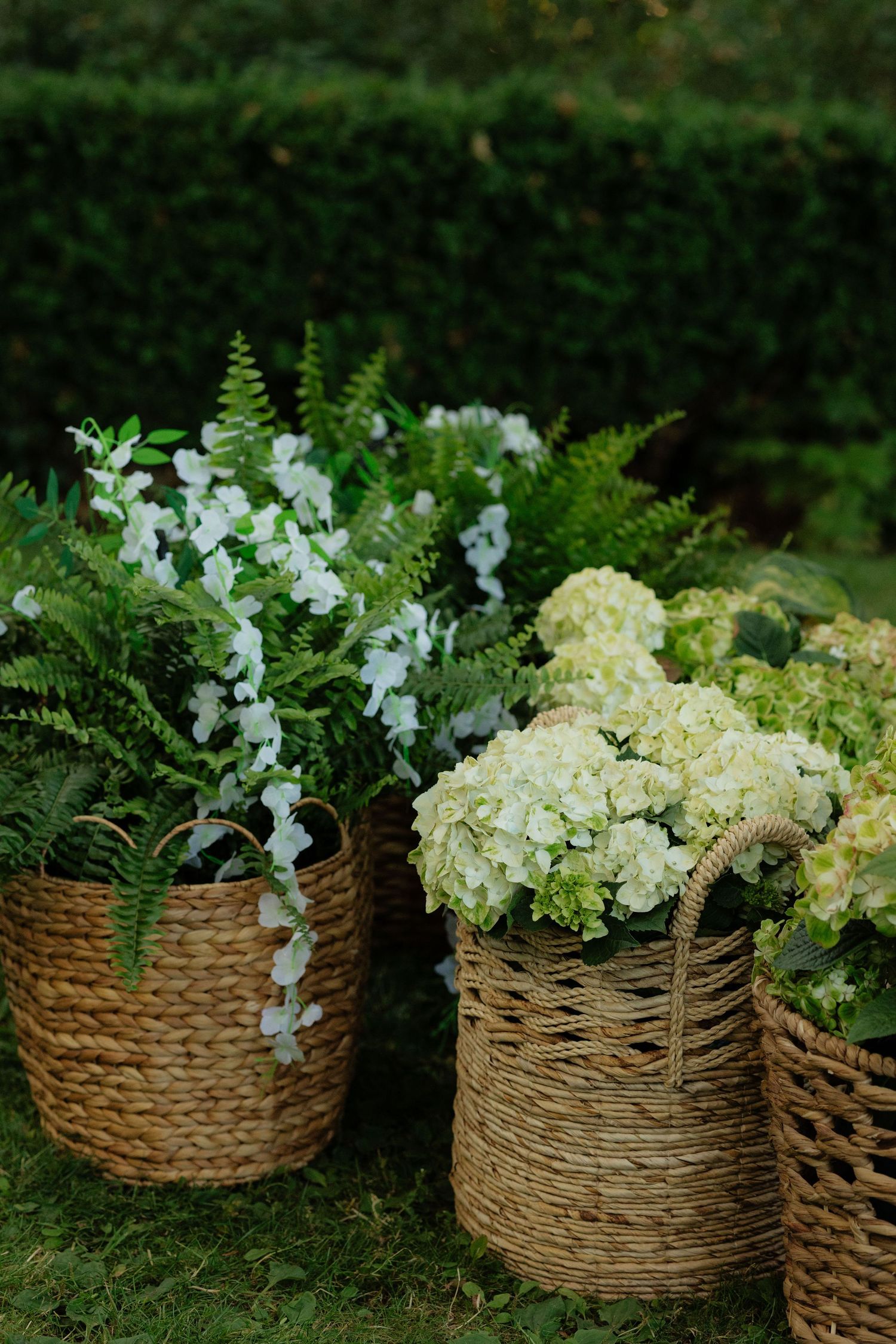 Three woven wicker baskets filled with white hydrangeas, ferns, and flowers on green grass at outdoor event.