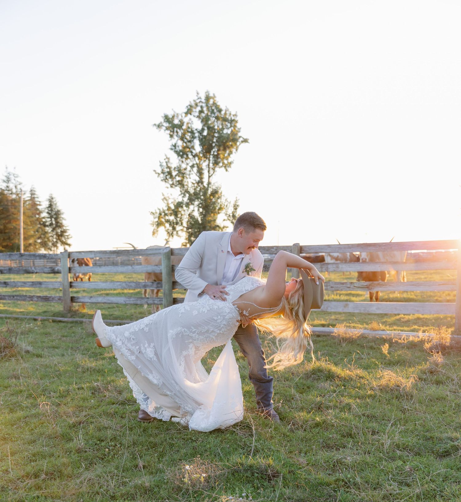 A couple in wedding attire dances in a grassy field at sunset with a wooden fence in the background.
